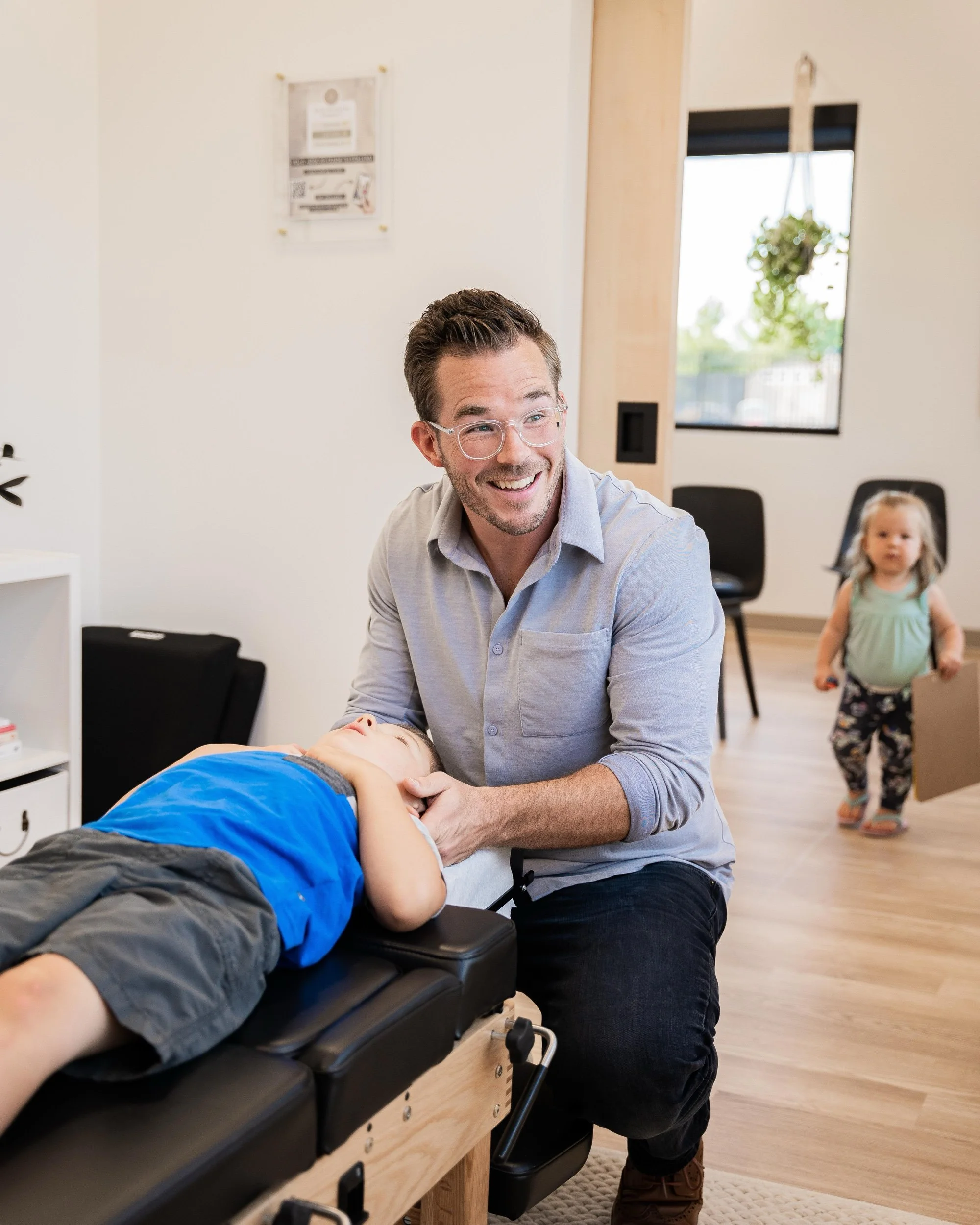 A man with glasses smiling while holding a young boy lying on a medical examination table in a clinic or therapy room, with a little girl standing in the background near a window.