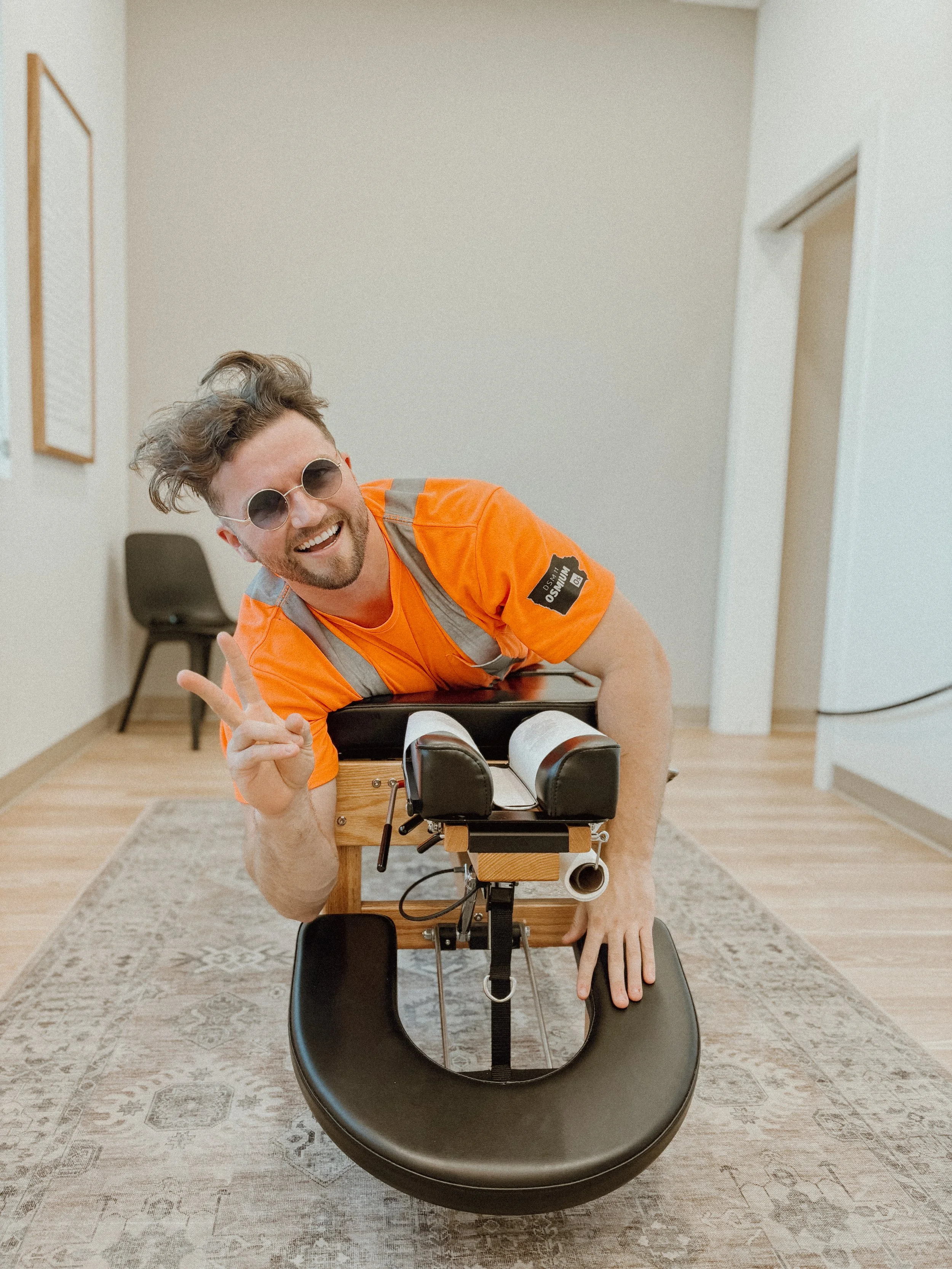 A man with curly hair and sunglasses making a peace sign, leaning on a chiropractic adjustment table in a room with beige walls and wooden flooring.