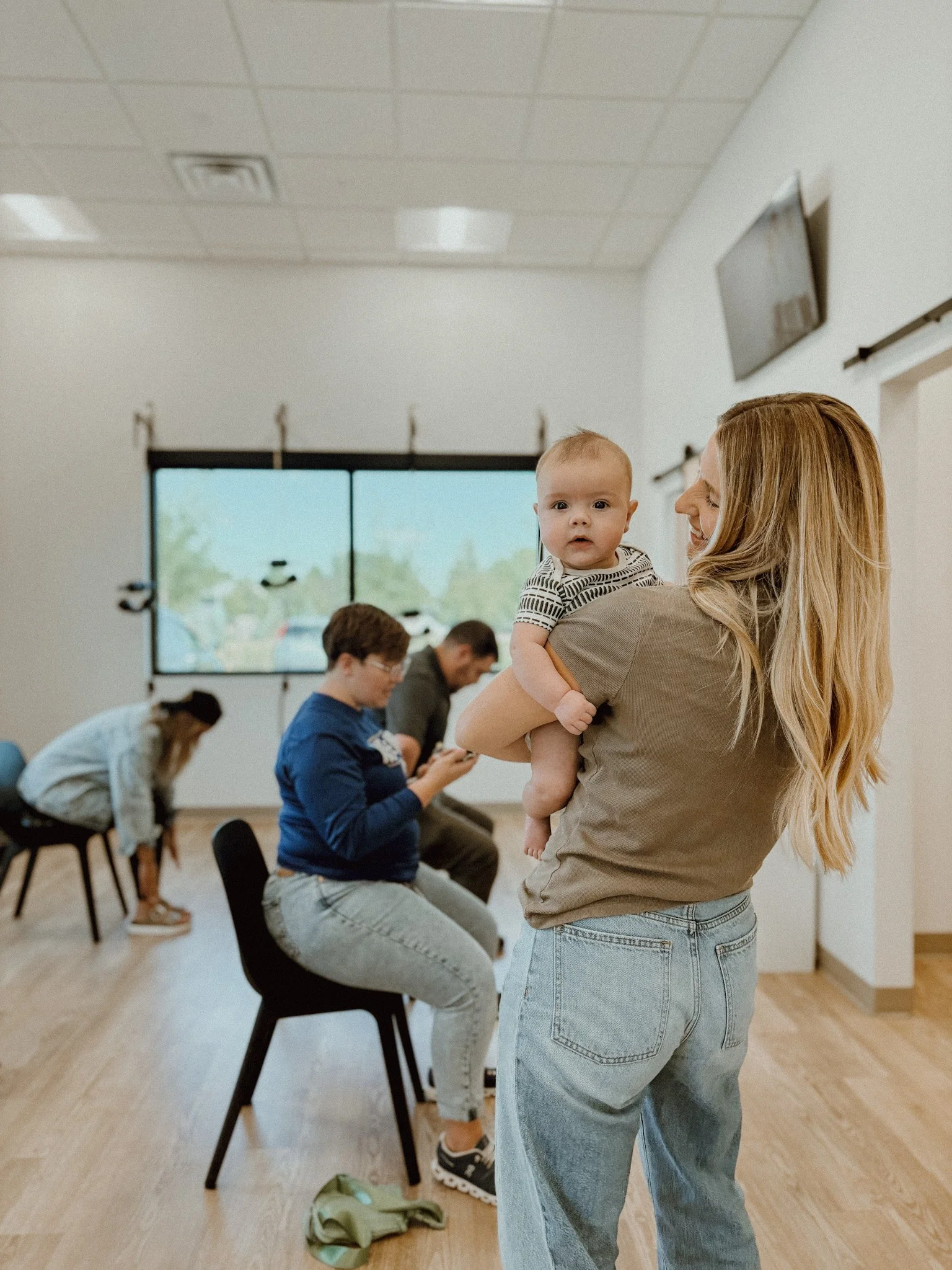 A woman holding a baby in a room with a wooden floor and a large window. Four people are seated in the background, two women and two men, using their phones or looking down. The room is well-lit with ceiling lights.