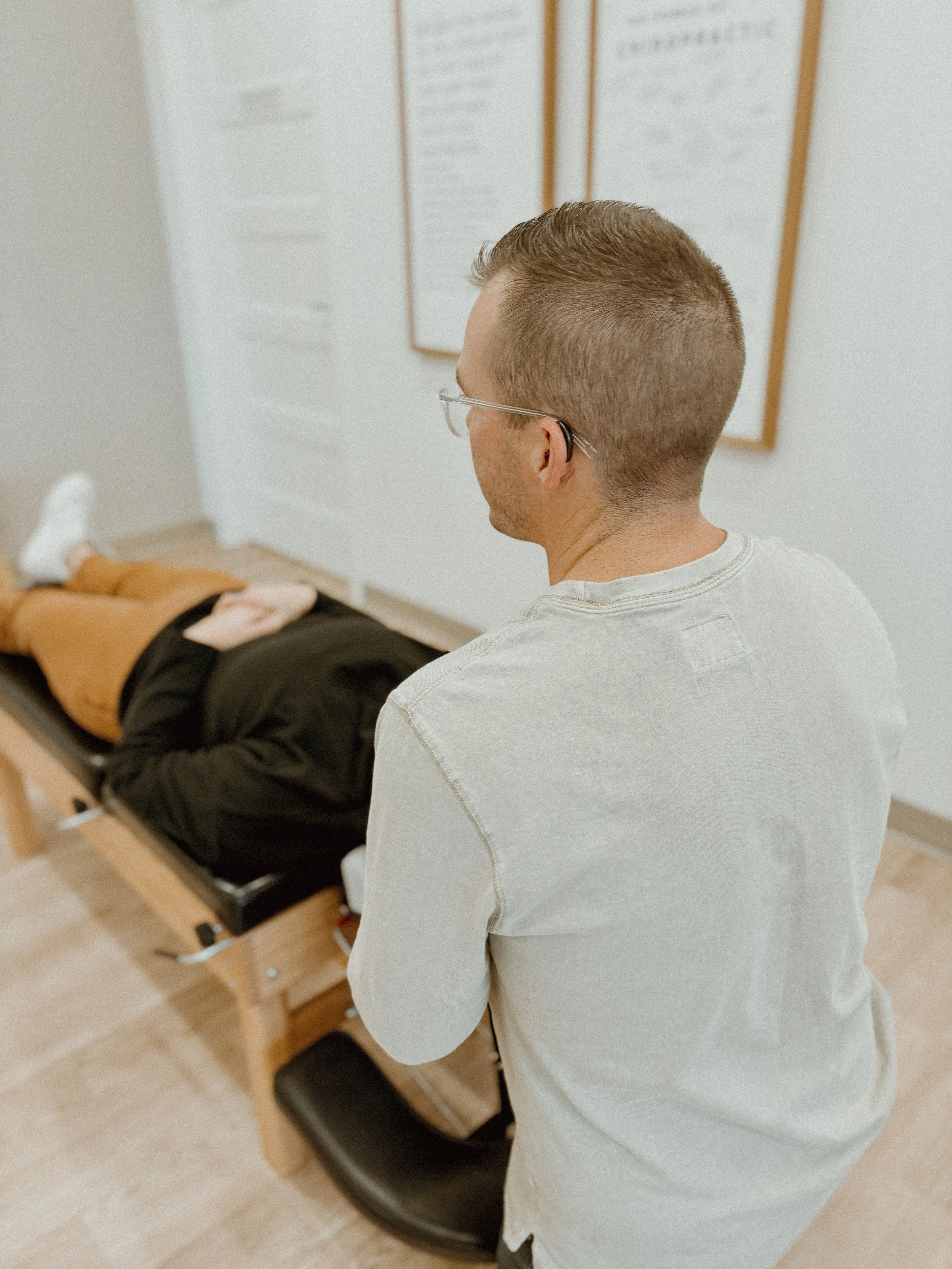 A man with glasses and a short haircut observing a woman lying on a medical examination table in a clinic or healthcare setting.