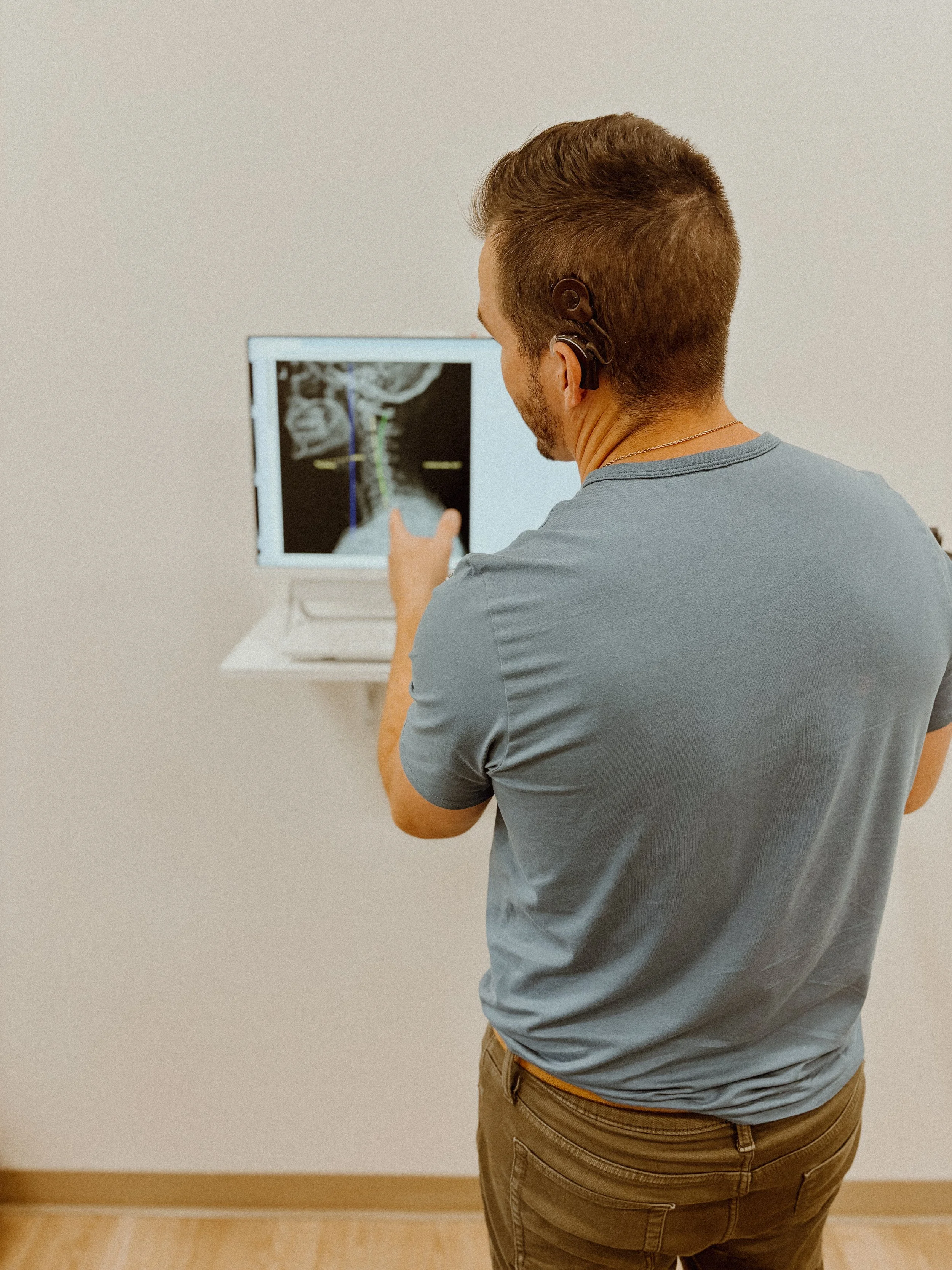 A man with short brown hair and wearing a blue t-shirt is examining a medical scan on a computer monitor, which shows an image of the neck and spine.