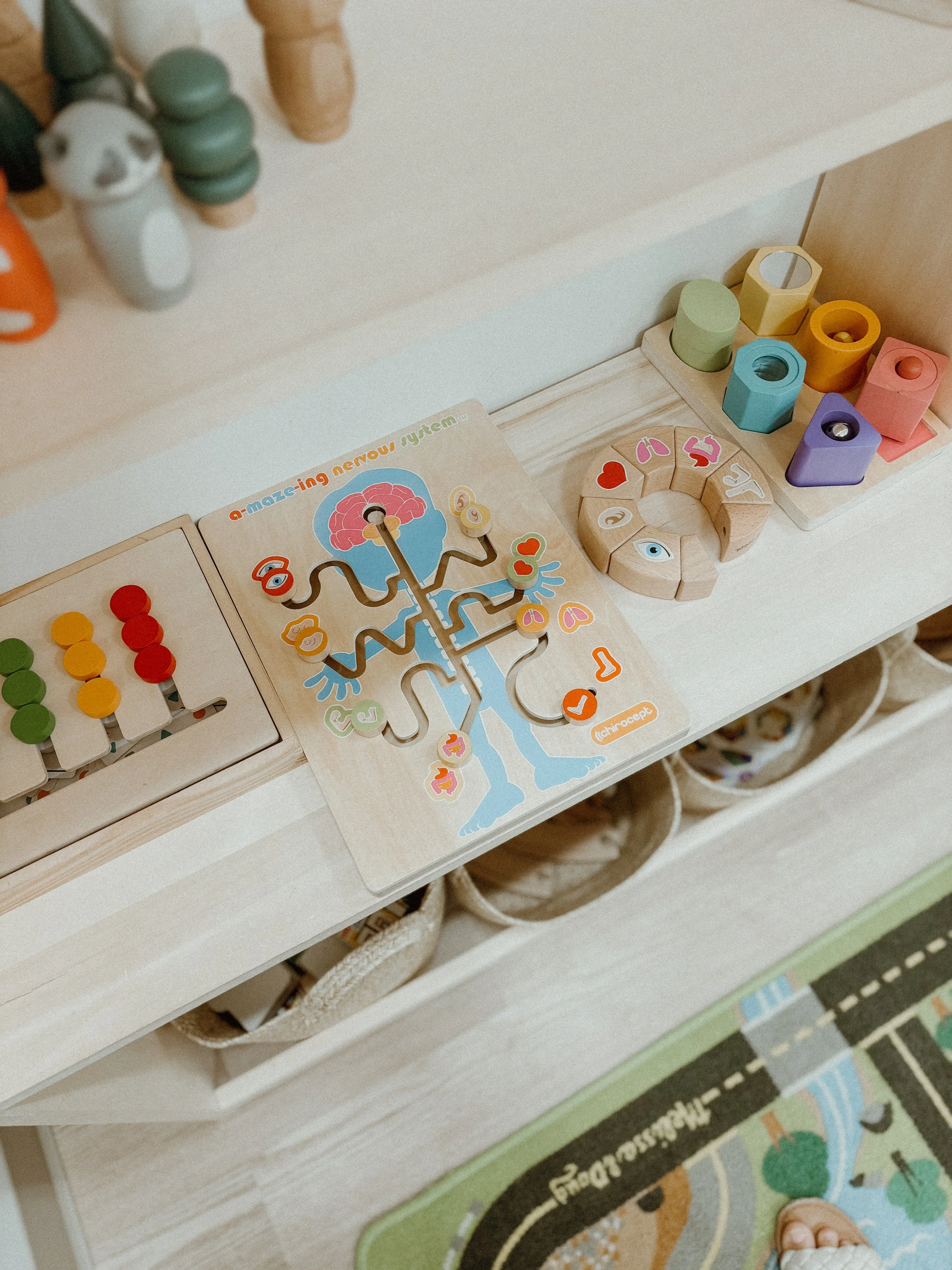 Children's toys on a white shelf, including a brain-themed maze game, multicolored wooden blocks, and a circular puzzle with animal and heart shapes, with part of a patterned rug and a child's toes visible in the corner.