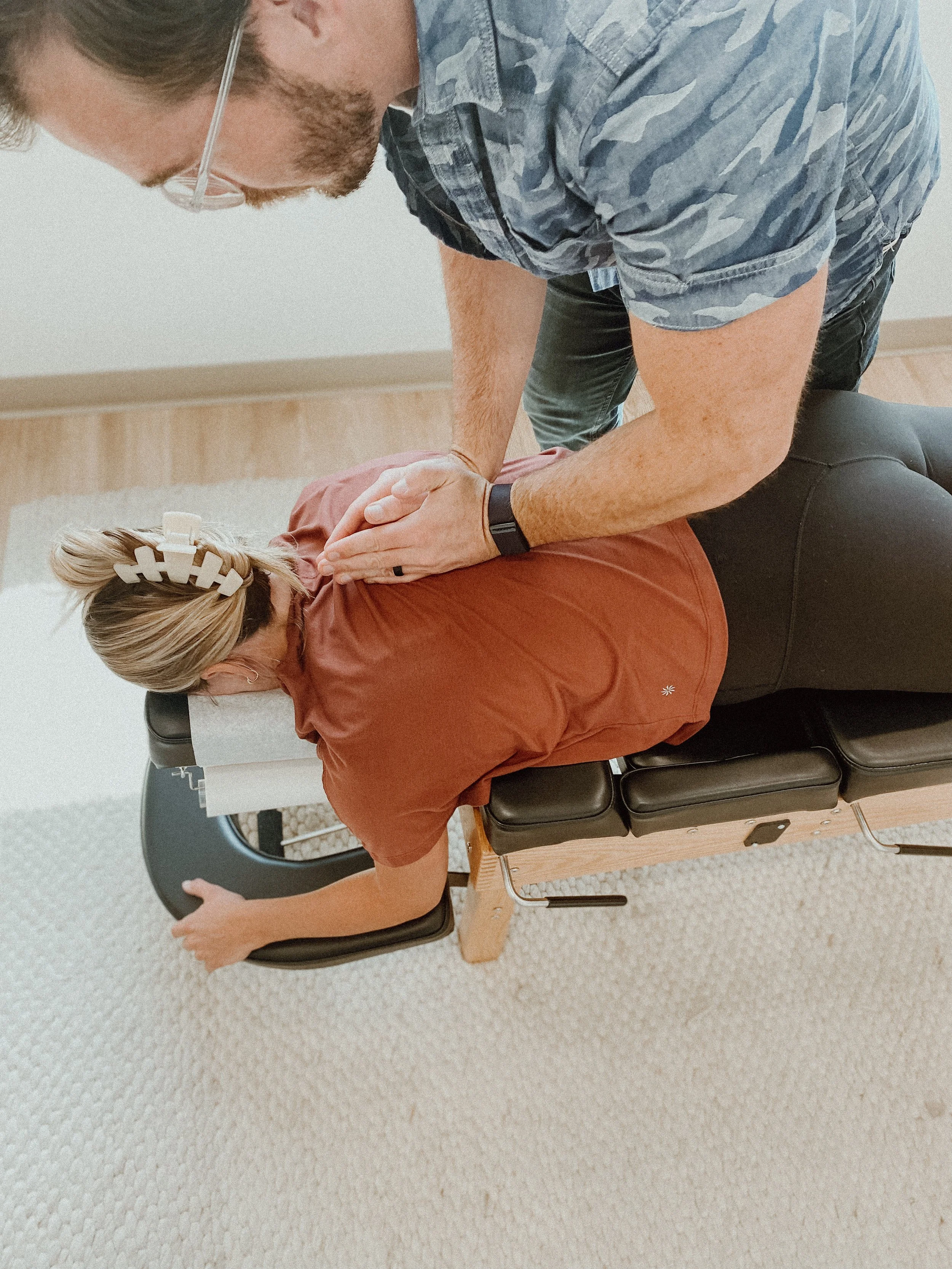 A woman lying face down on a chiropractic or physical therapy table receiving a back adjustment from a male practitioner.