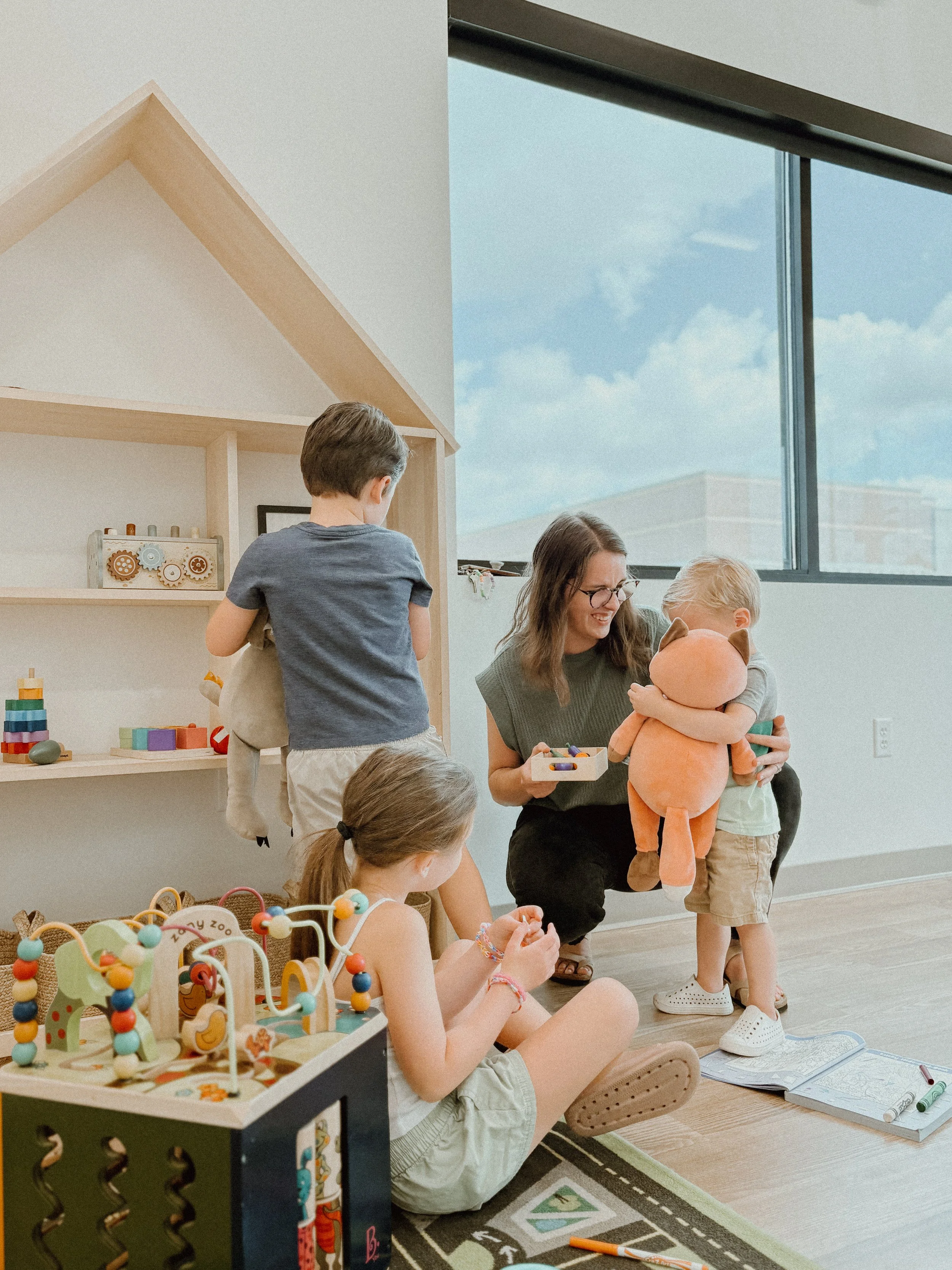 A woman is holding a large plush doll while smiling at a young boy hugging the doll. Two children are sitting on the floor with toys, and another child stands nearby, all in a playroom with toys and a house-shaped shelf.