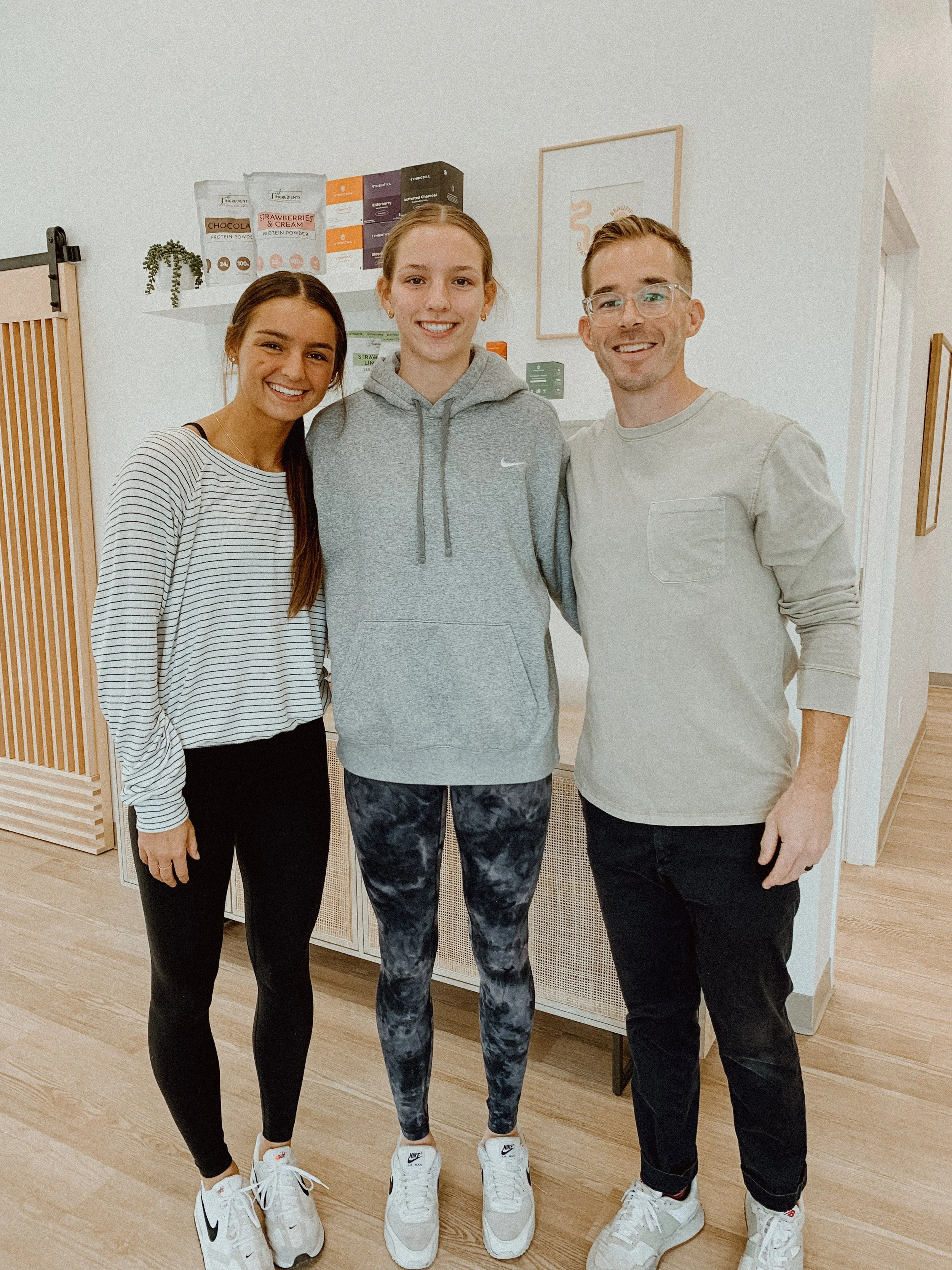 Three people standing inside a room, smiling at the camera. The person on the left is a young woman with long dark hair, wearing a striped long sleeve top, black leggings, and white sneakers. The person in the middle is a young woman with red hair, wearing a gray Nike hoodie, patterned leggings, and white sneakers. The person on the right is a young man with short blonde hair and glasses, wearing a light gray long sleeve shirt, black pants, and white sneakers.