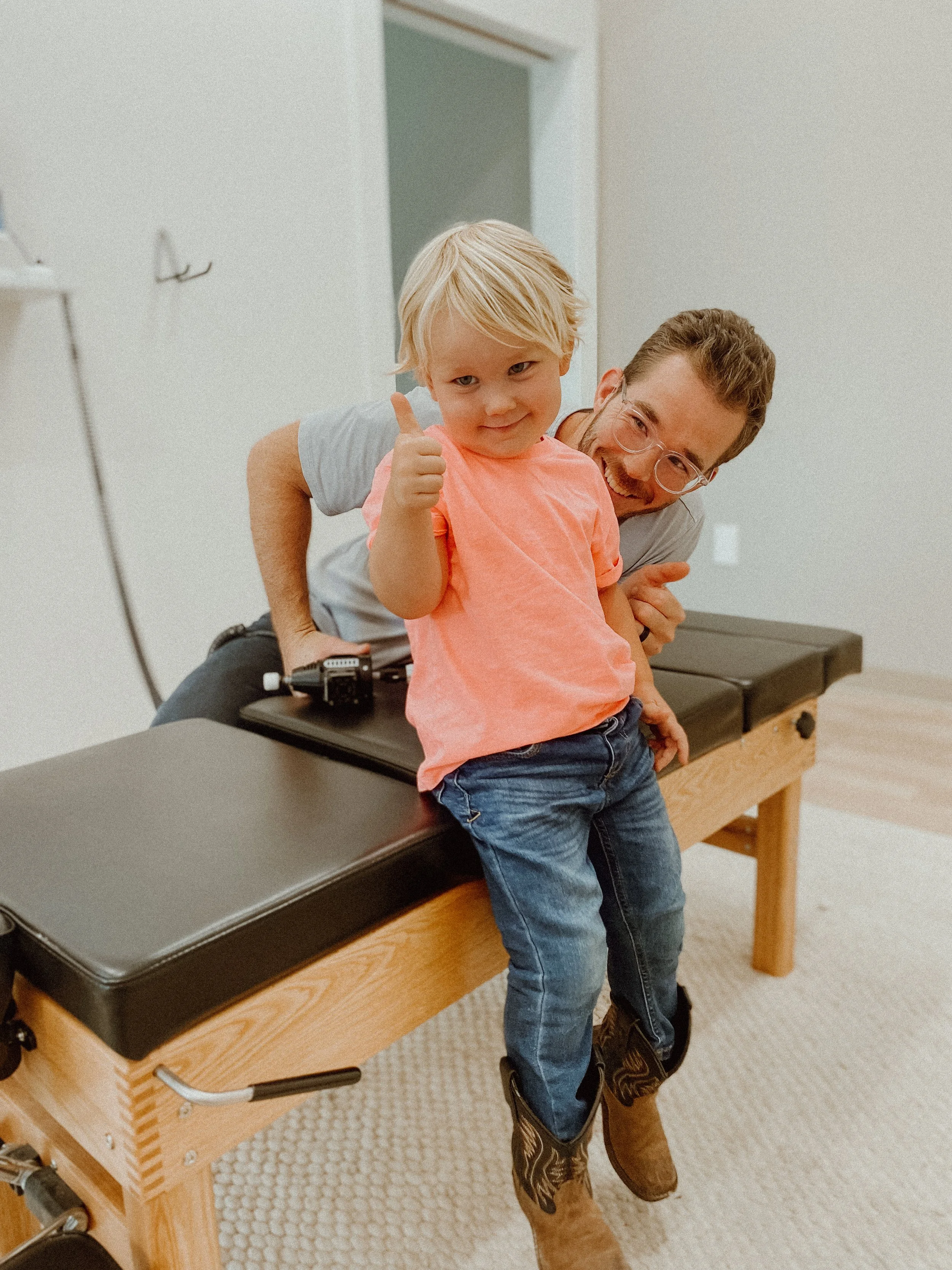 A young boy sitting on an examination table at a doctor's office, giving a thumbs-up, with a smiling healthcare professional in the background.
