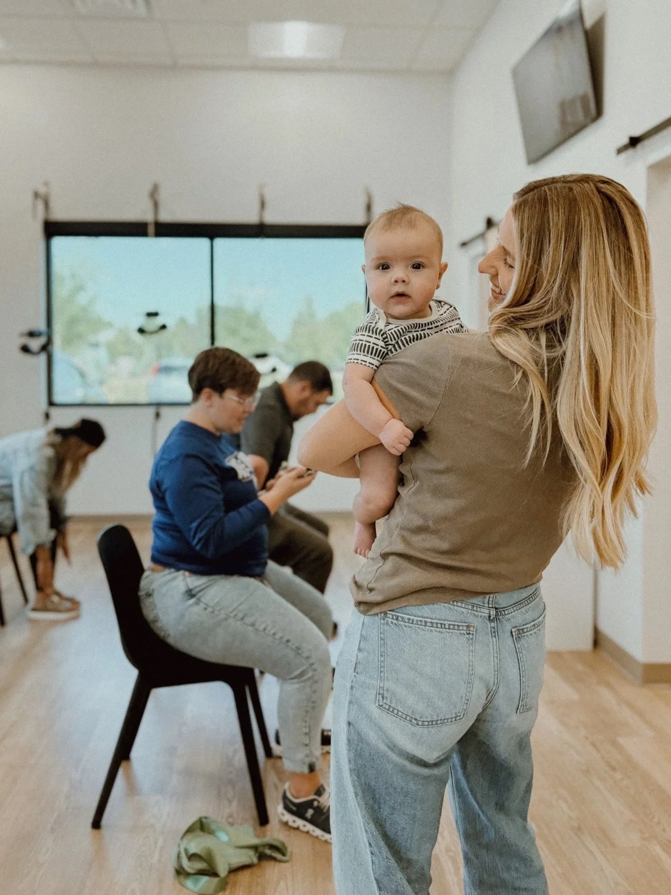 A woman holding a baby in a room with large windows and several people sitting and standing, some using phones.