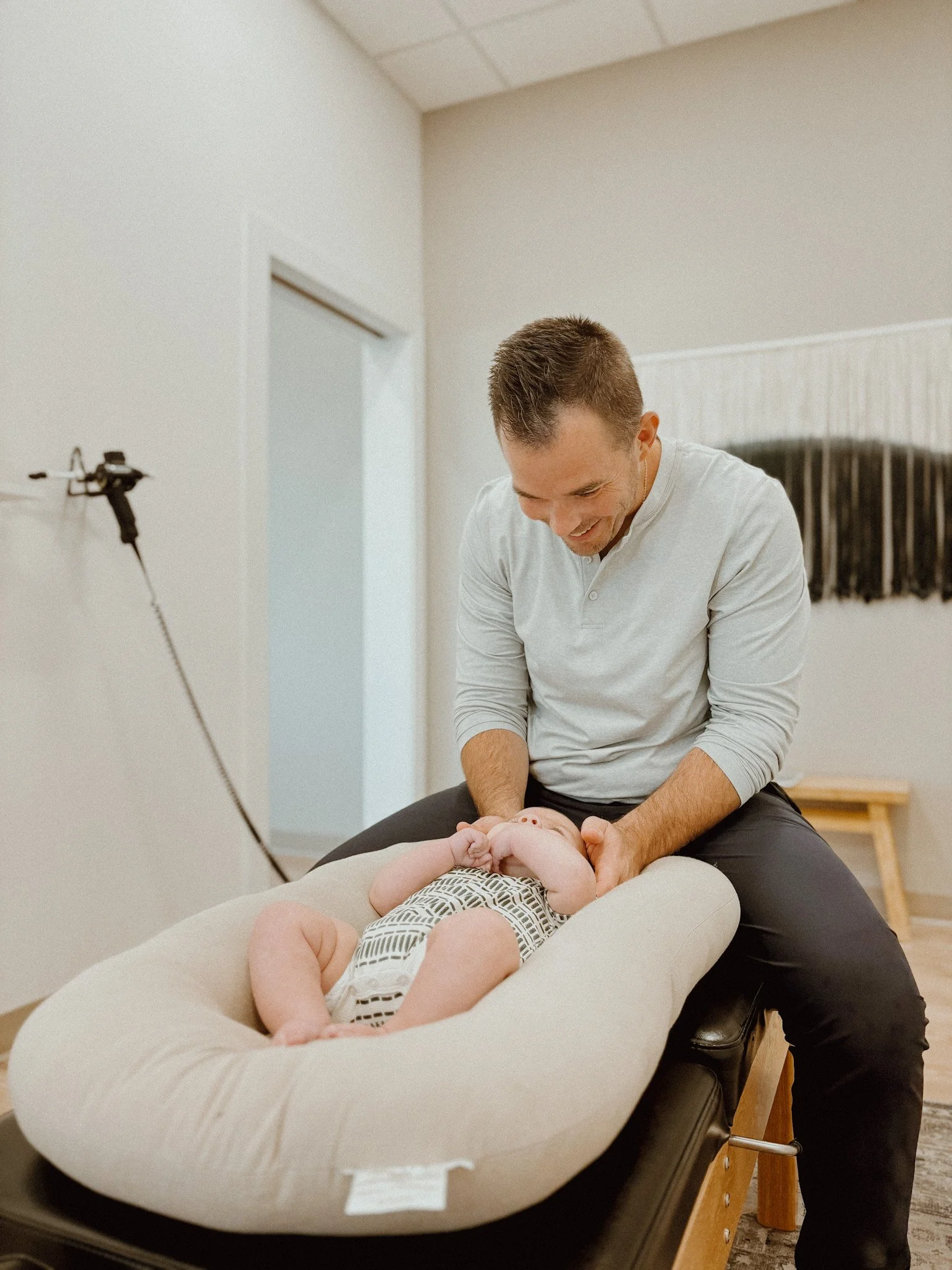 A man smiling and looking down at a baby lying on a cushioned changing pad. The man is seated, and the baby is wearing patterned clothing. The room has beige and white walls with minimal decor.