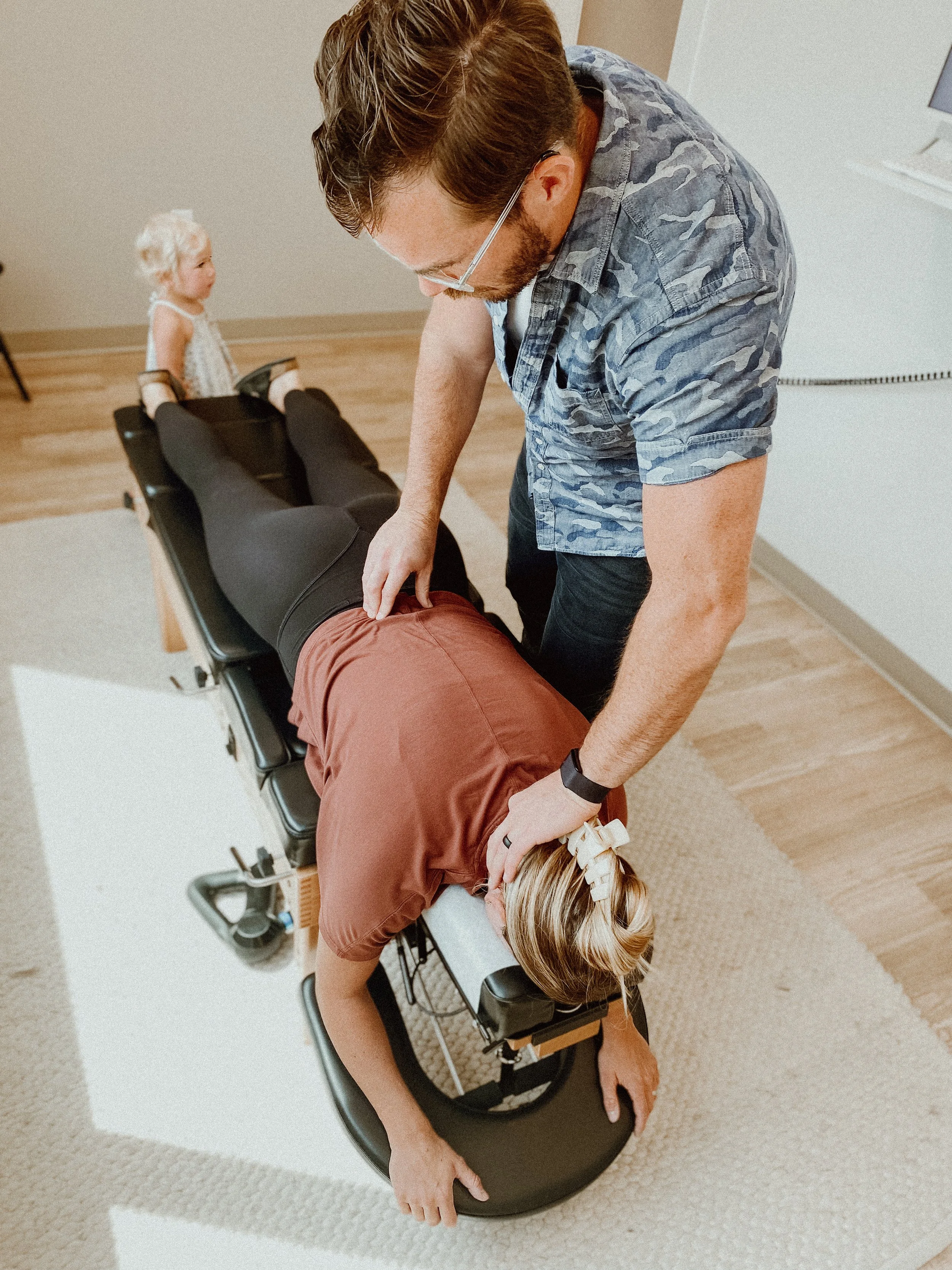 A chiropractor adjusting a woman lying face down on a chiropractic table, with a young girl standing in the background, observing.