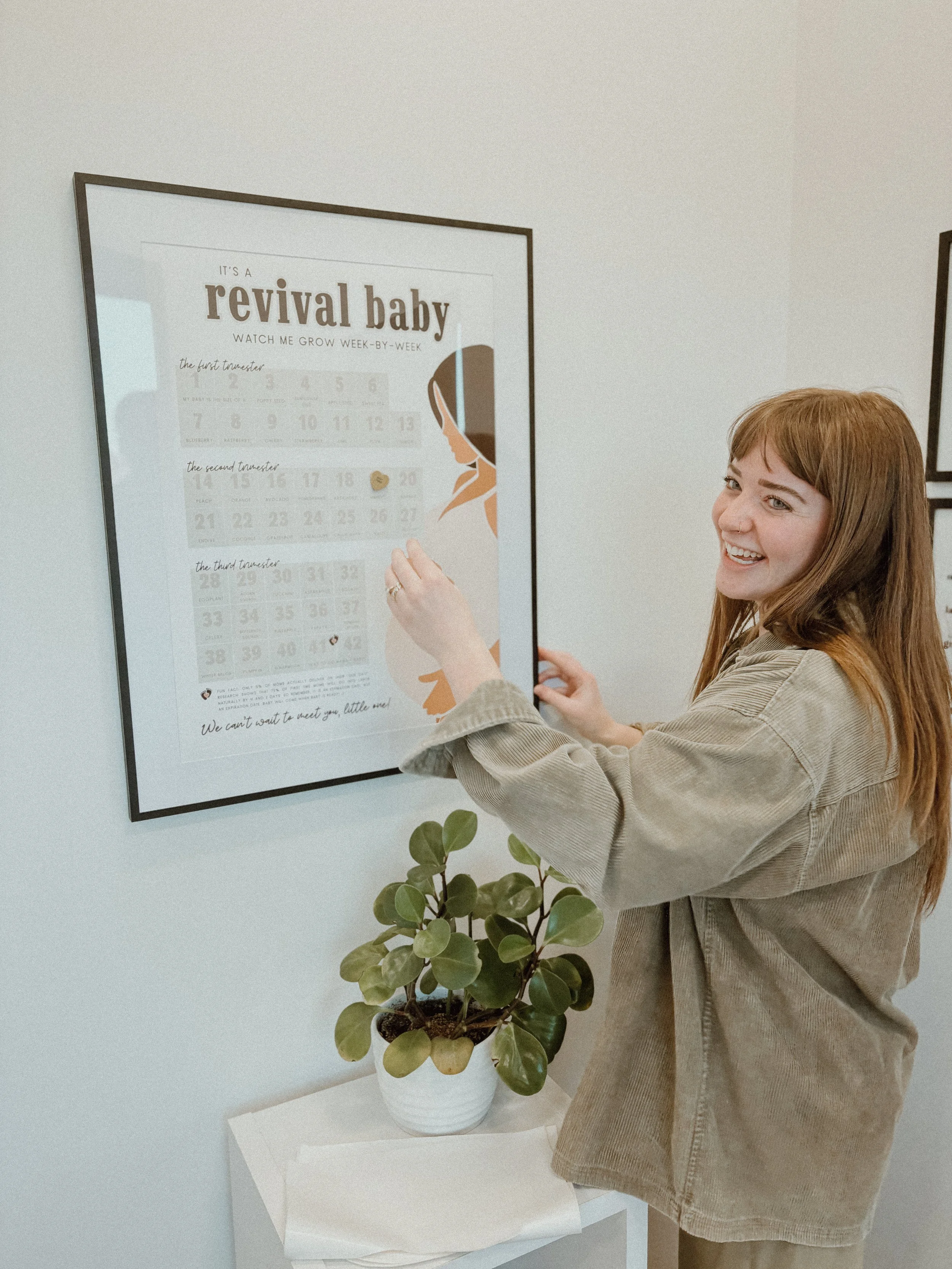 A woman with red hair smiling as she points at a large framed milestone poster for a baby's development on the wall, with a potted plant on a small table below.
