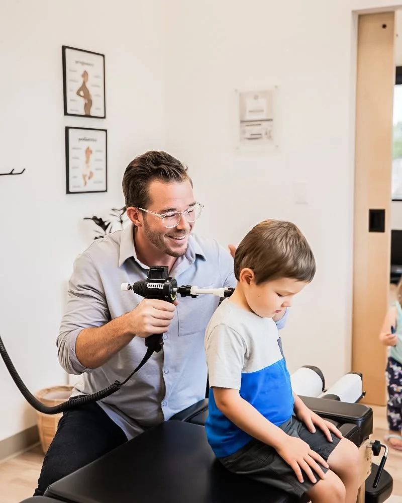 A male doctor examines a young boy's ear with an otoscope in a medical office, with the boy sitting on an exam table.