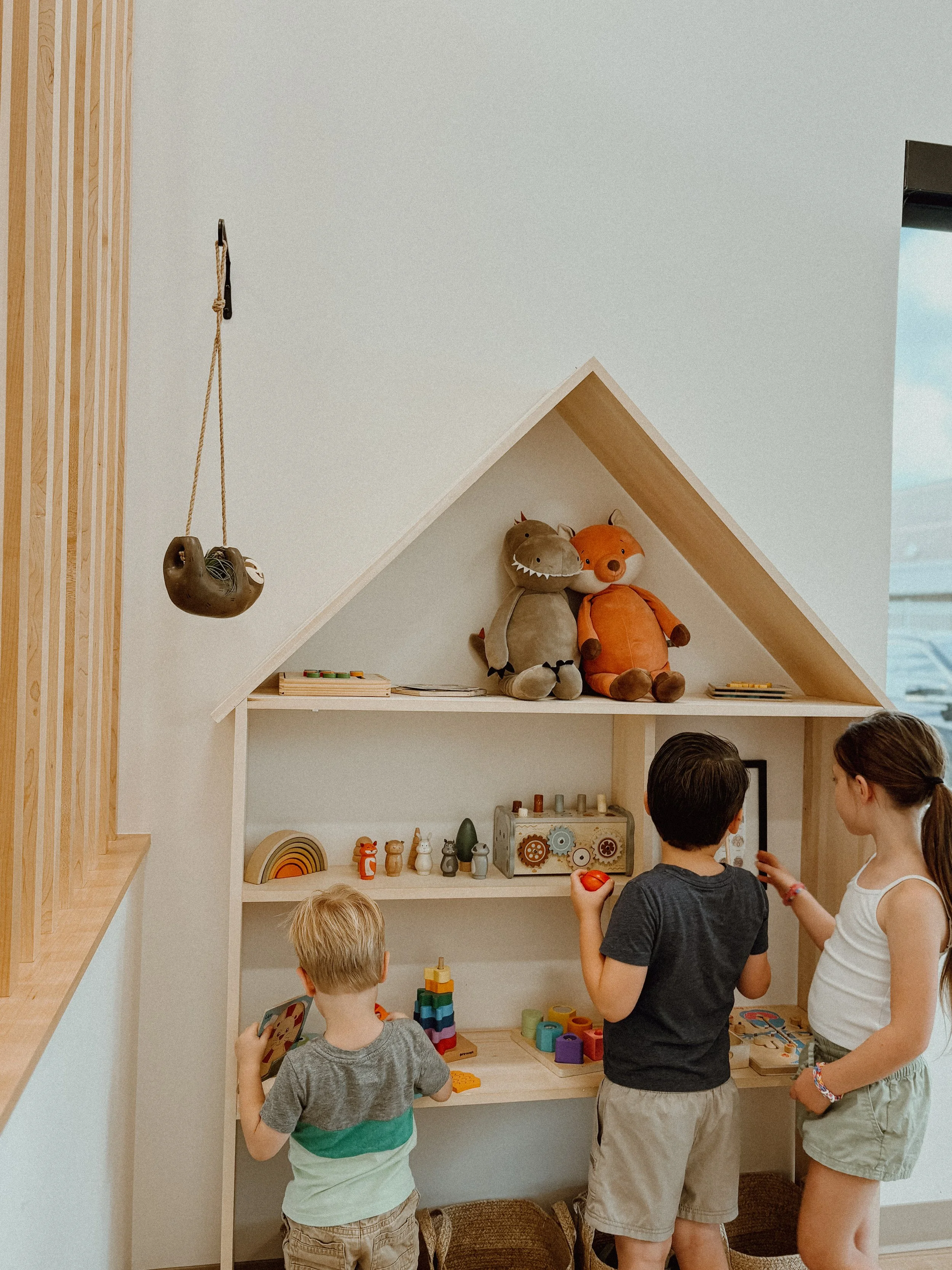 Three children are playing with toys in front of a wooden playhouse shelf in a children's playroom.