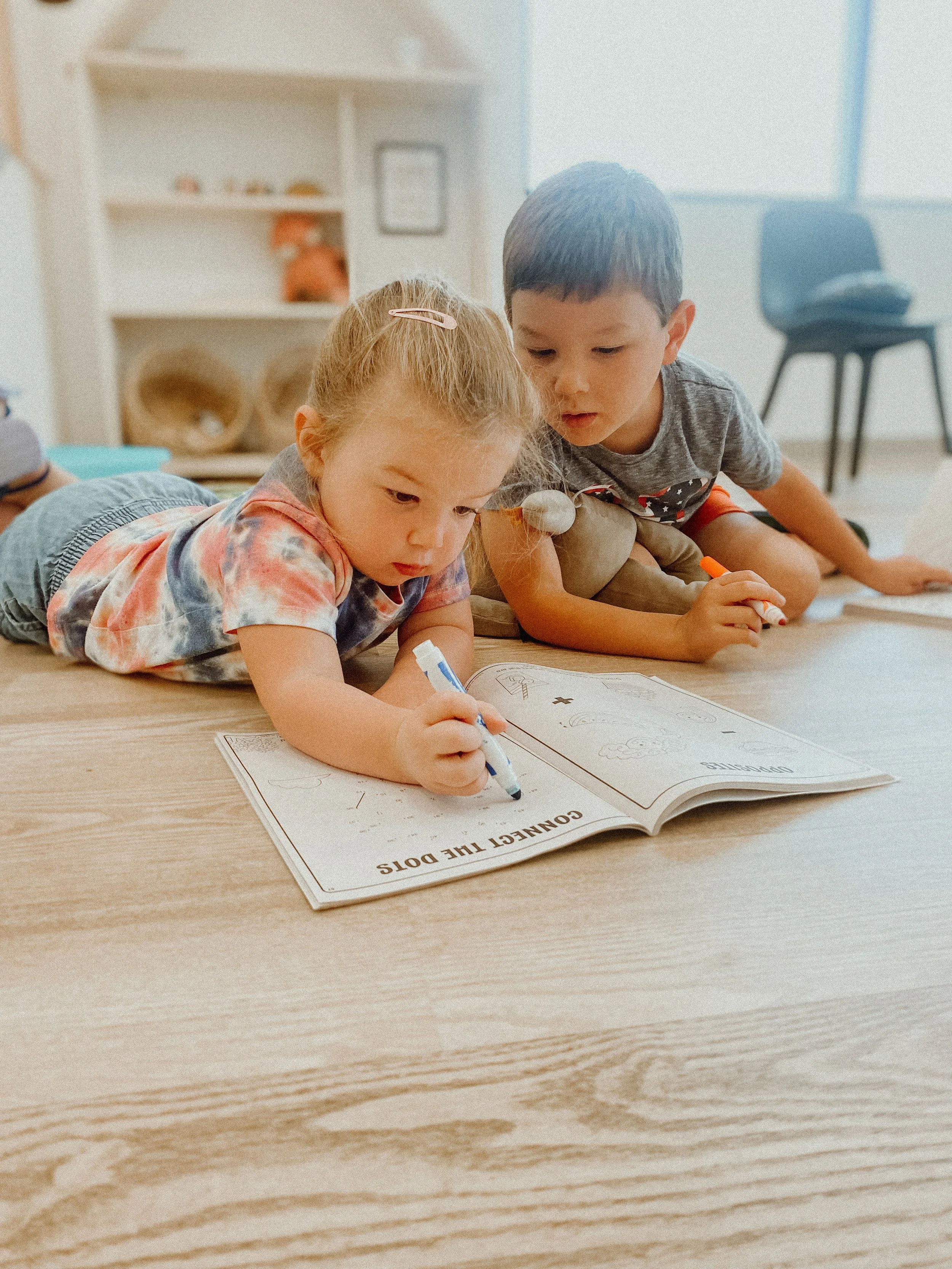 Two young children lying on the floor, coloring in a workbook titled 'Connect the Dots'.