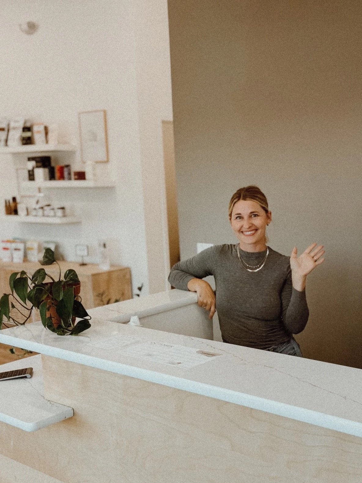 A smiling woman in a gray long-sleeve shirt sitting at a counter, waving. The background includes white shelves with jars and books, a wooden cabinet, and a plant on the counter.