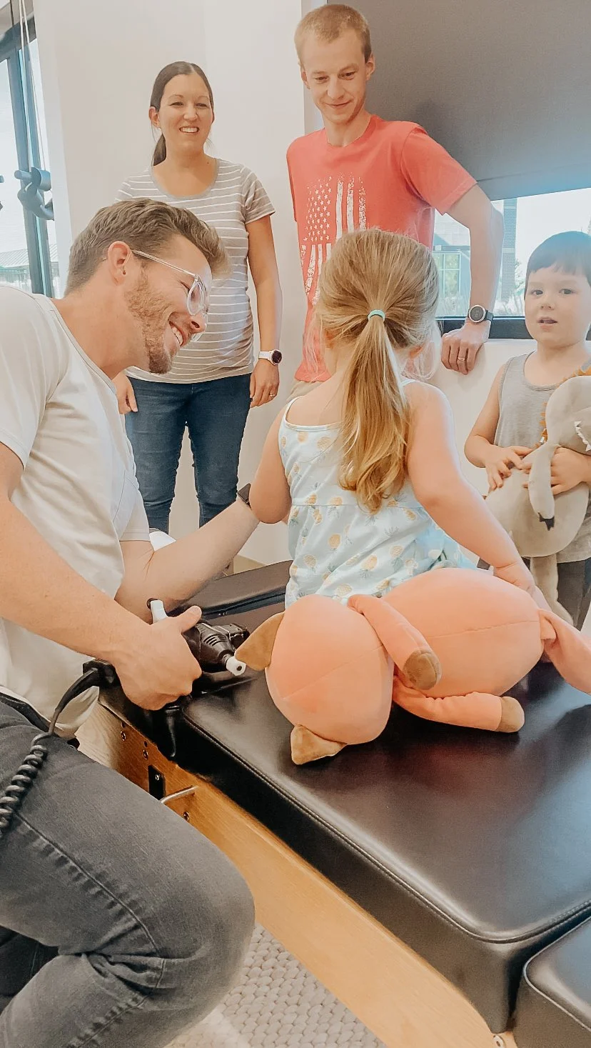 A young girl with a ponytail sitting on a medical examination table, being examined by a male medical professional while family members watch in a bright room.
