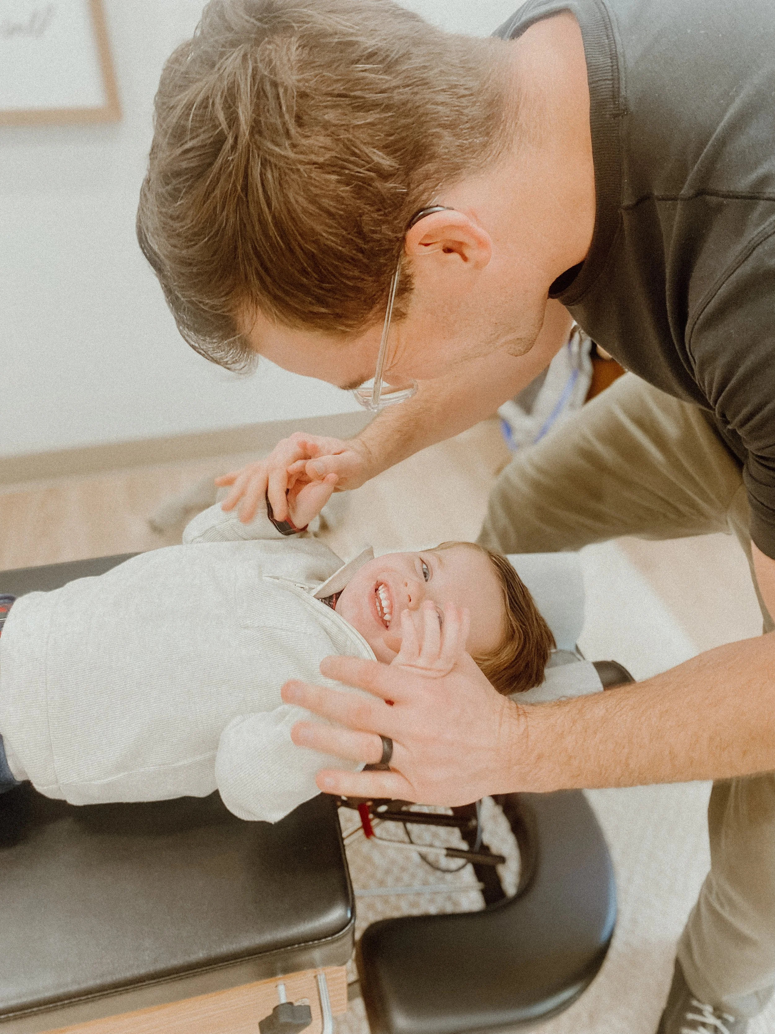 A young boy lying on an examination table, smiling as a dentist or doctor checks his teeth, with both appearing happy.