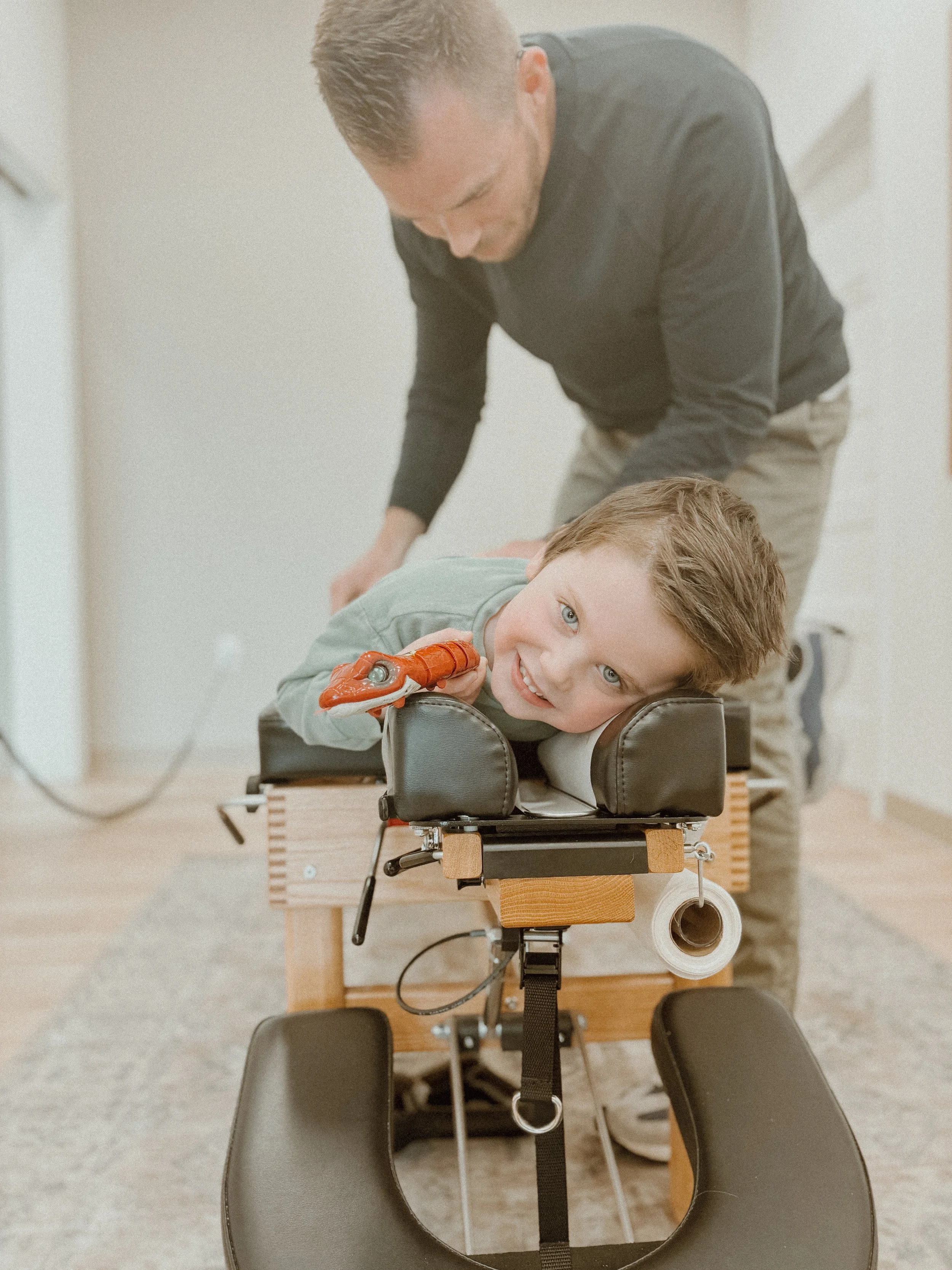 A young boy smiles while lying face down on a chiropractic adjustment table, with a person standing behind him preparing for a chiropractic treatment in a clinic.