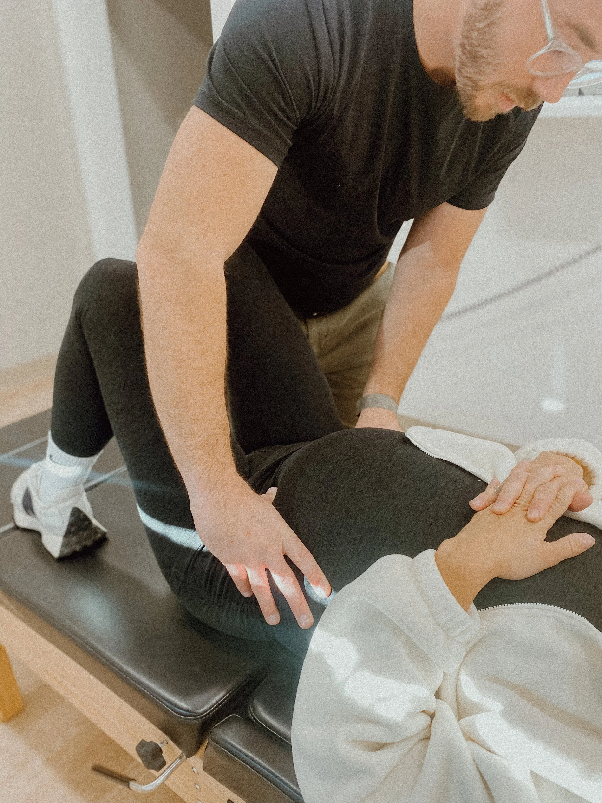 A person in a white hoodie is lying face down on a black massage or chiropractic table, while another person in black clothing is assisting or performing a procedure on their knee or thigh.