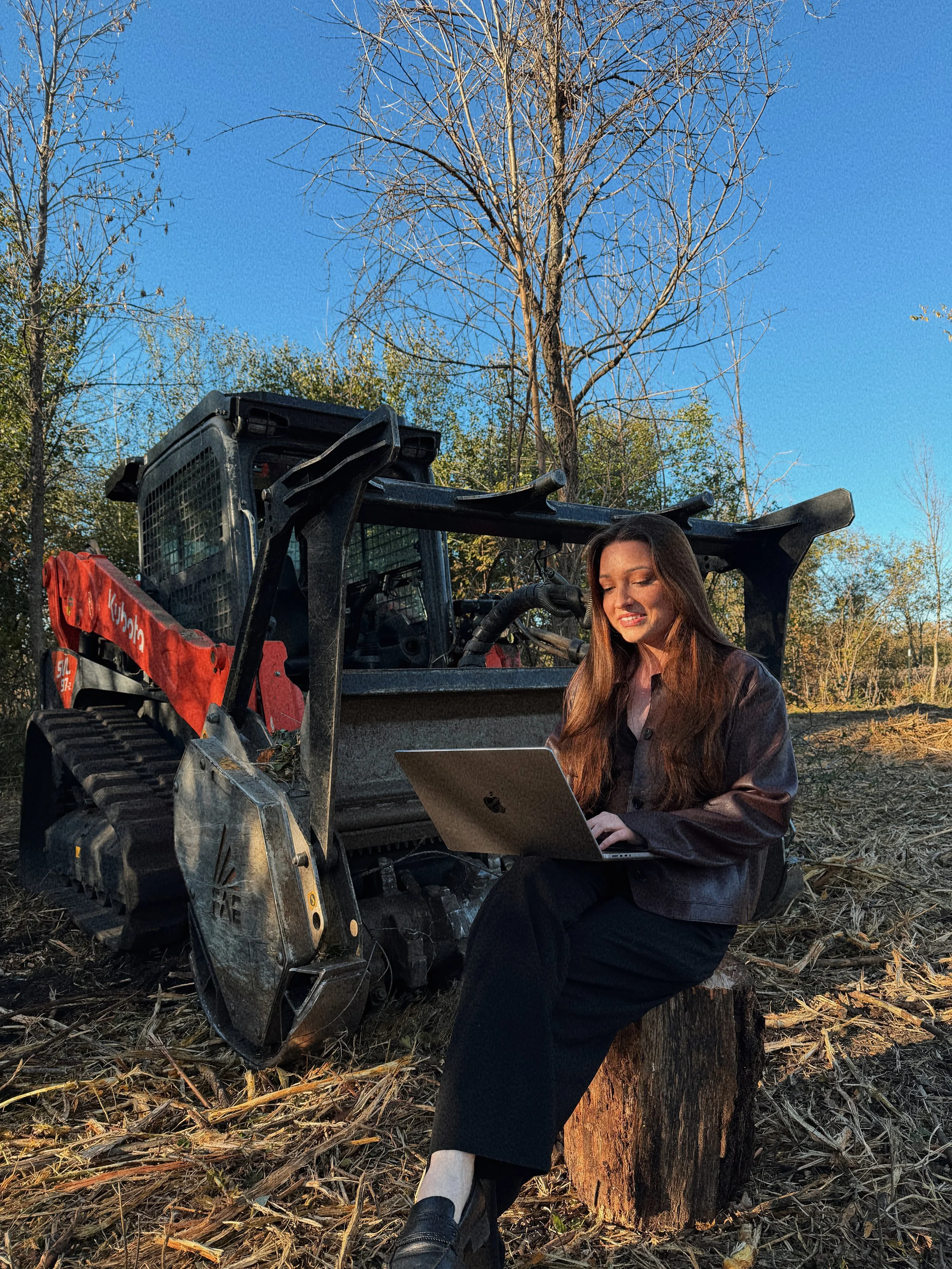 A woman sitting on a tree stump using a laptop outdoors near a Bobcat tractor, with trees and clear blue sky in the background.