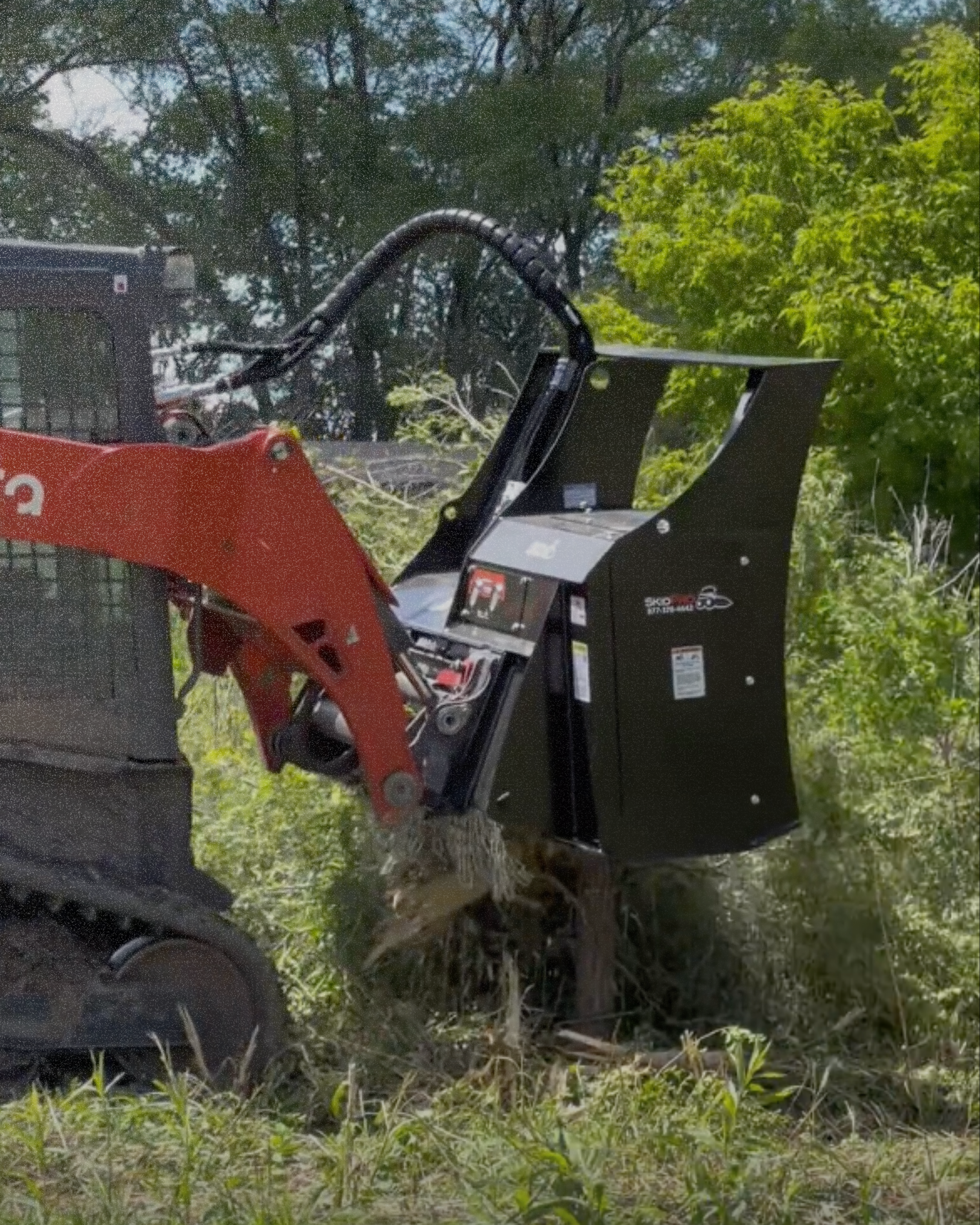 A small excavator with a black attachment clearing brush and small trees in a green outdoor area.