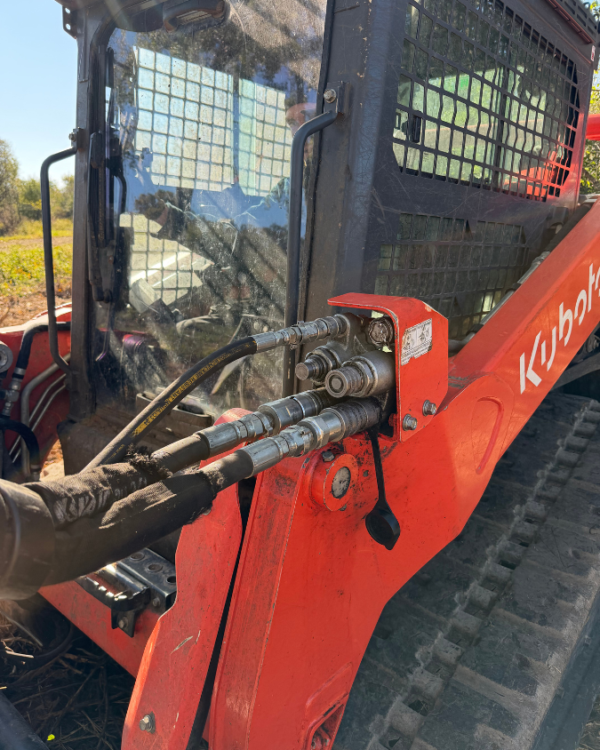 Close-up of an orange Kubota skid steer loader with hydraulic hoses connected to the lift arm, viewed from the side with a partially visible protective black cage and window.