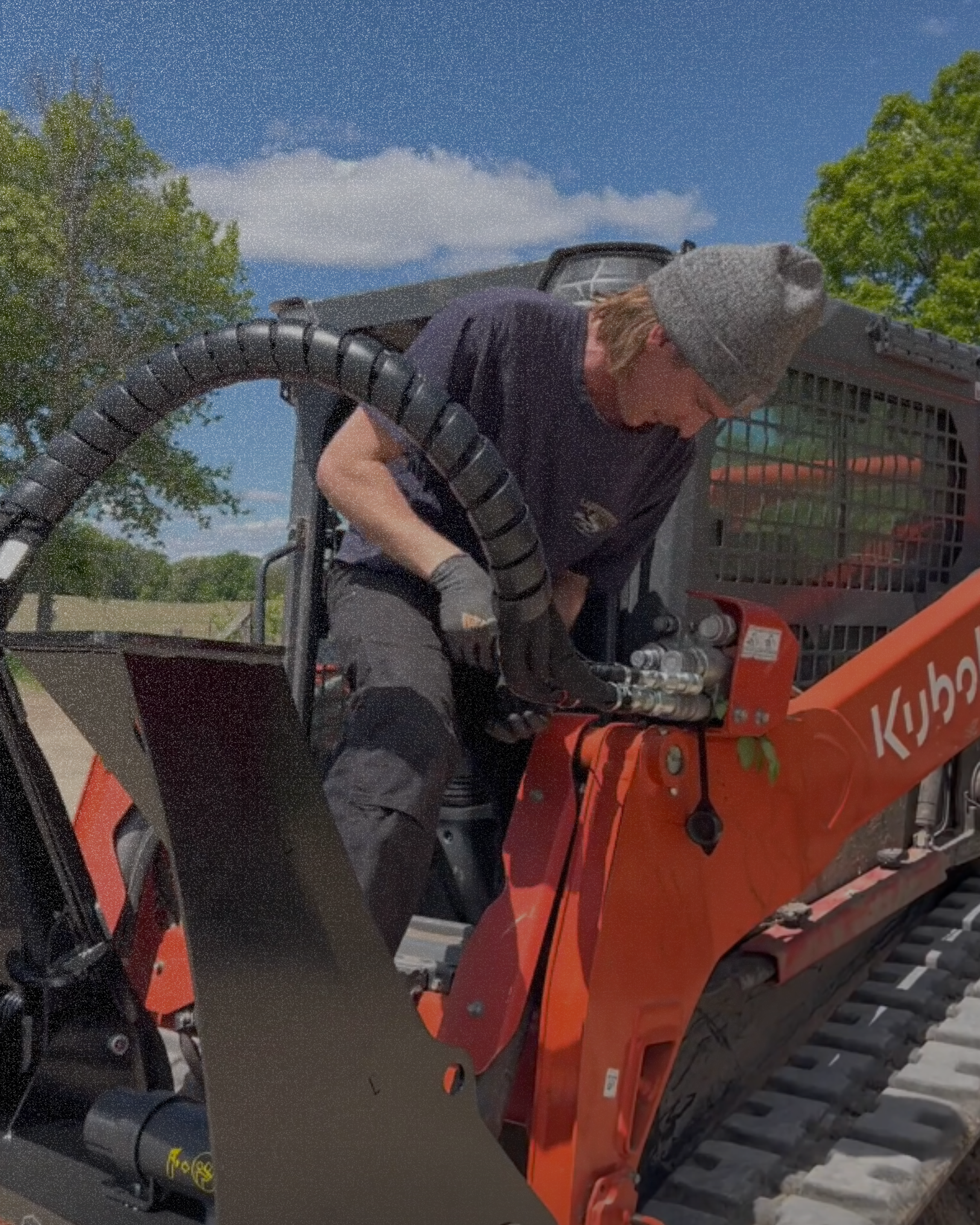 A person operating a Kubota compact track loader outdoors, wearing a gray knit hat, black gloves, and a black shirt while working on hydraulic hoses.