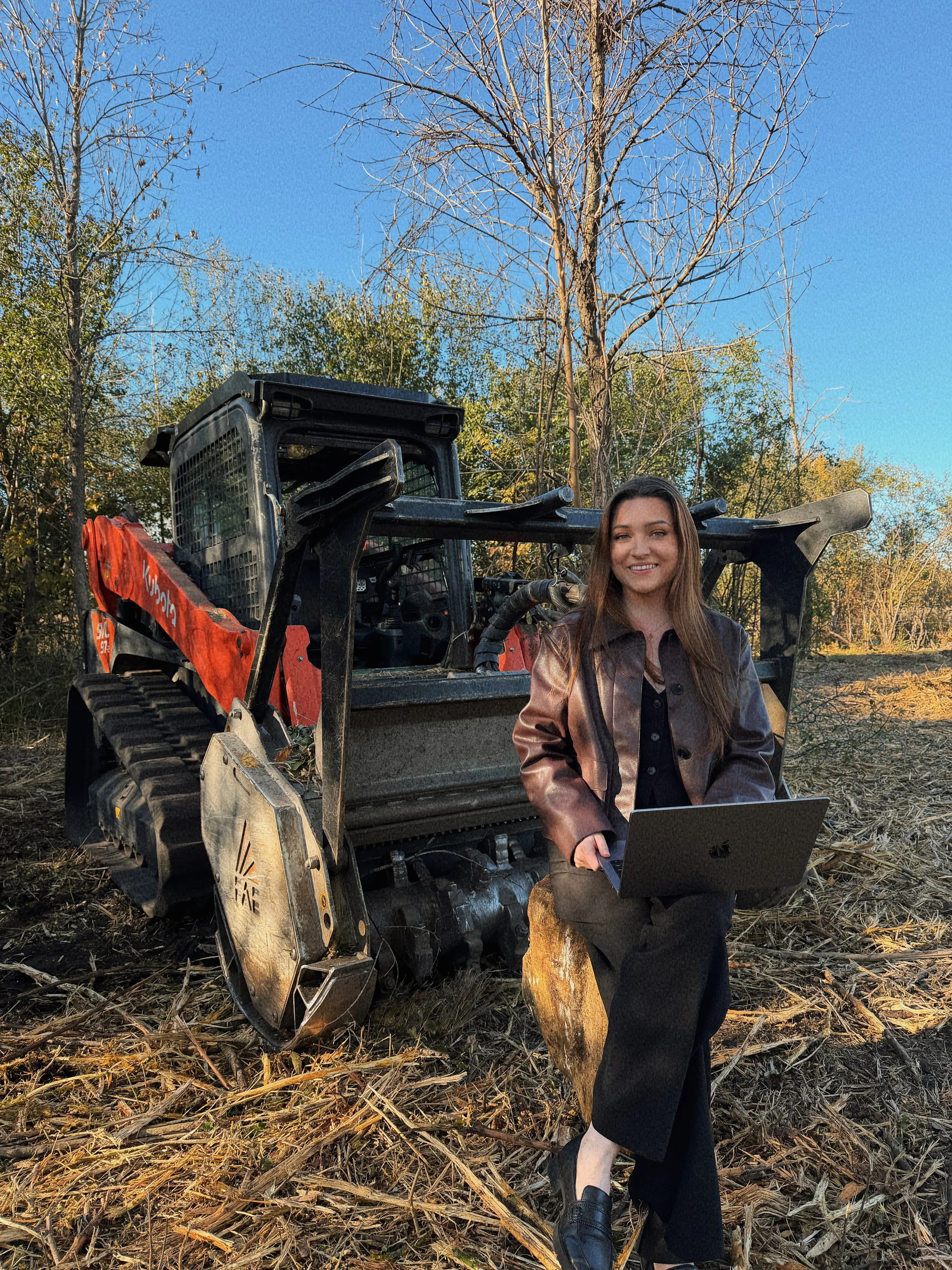 A woman standing outdoors in a cleared wooded area with trees and a blue sky, smiling while holding a laptop, next to a small urban excavator.