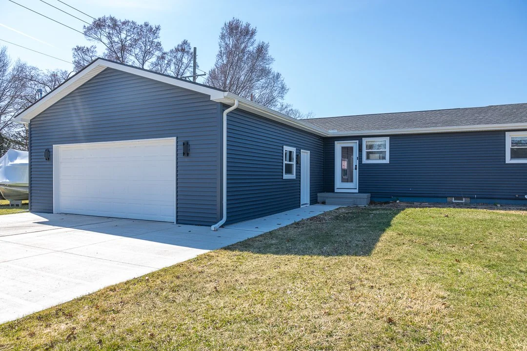A house with blue siding, a white garage door, and a small front porch, with a lawn and trees in the background.