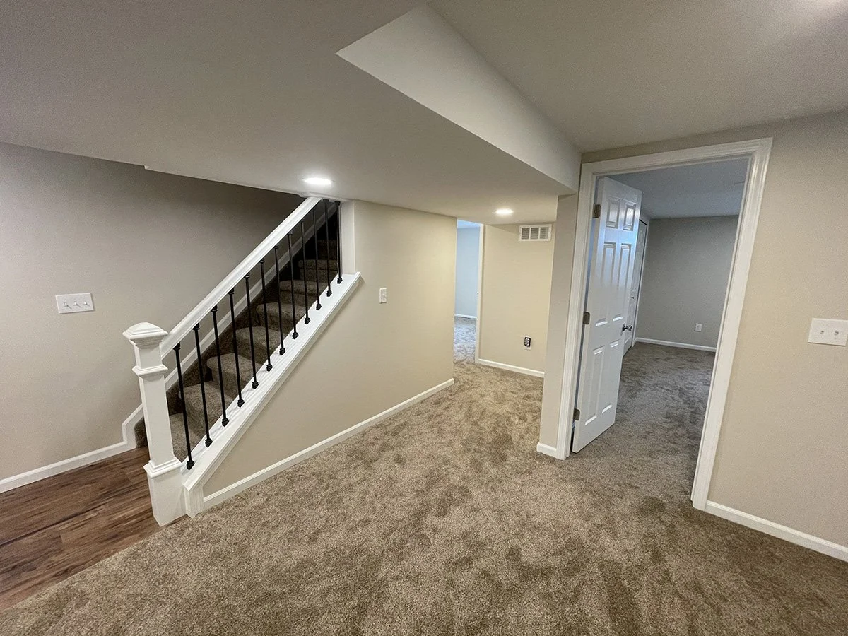 Interior of a house showing a staircase with carpeted steps, beige walls, white trim, open door to a room with carpet, and a hallway with carpeted floor.