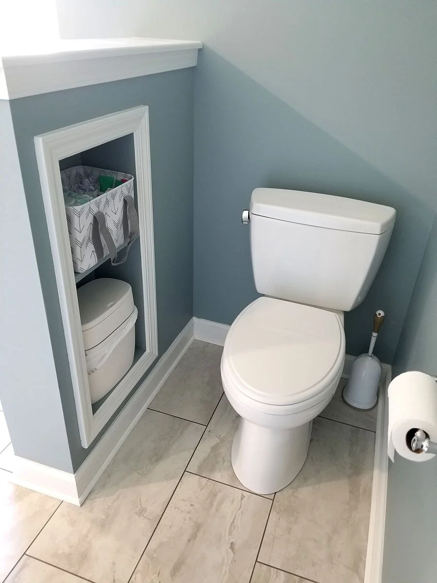Small bathroom with a white toilet, a wall-mounted toilet paper roll, a corner with an access panel containing a waste bin, and a toilet brush, with light-colored tiled flooring and gray-blue colored walls.