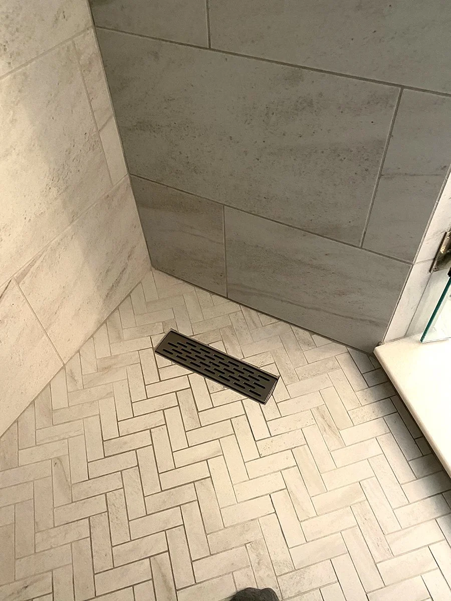 Floor drain in a bathroom shower with herringbone-patterned tile and a glass shower wall.