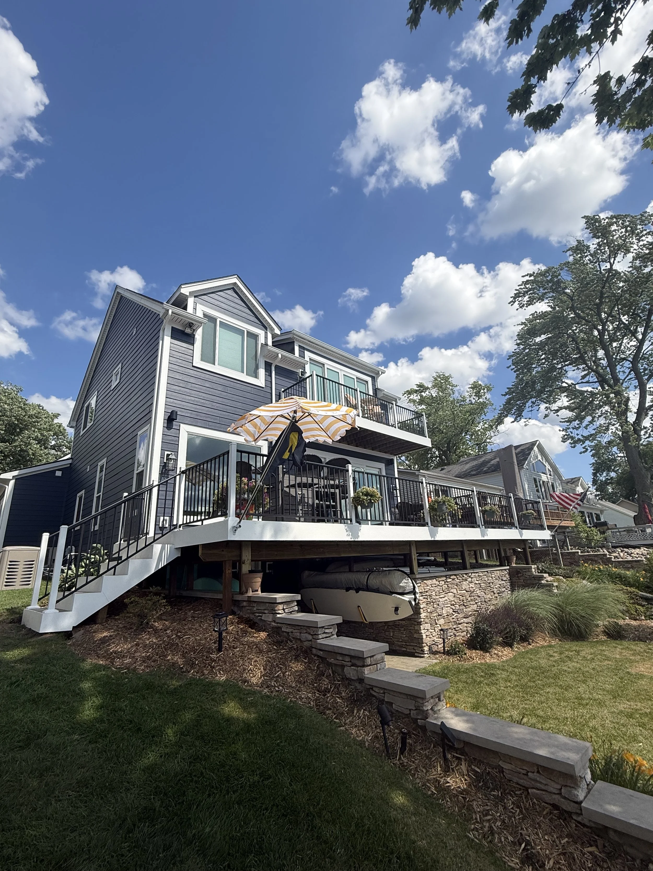 A multi-story house with gray siding, white trim, and a large wooden deck with black railing, decorated with potted plants and outdoor furniture, situated under a partly cloudy blue sky and surrounded by trees and landscaping.