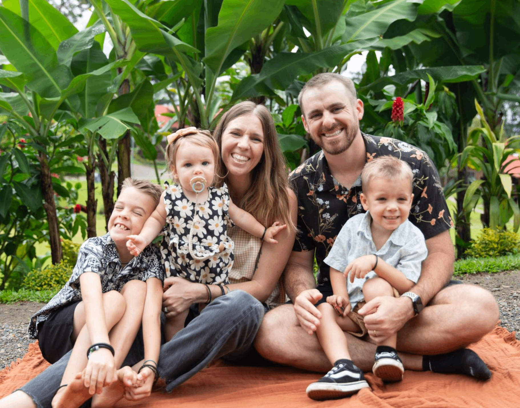 A family of five sitting on a blanket outdoors surrounded by large green plants and corn stalks, smiling and posing for the photo.