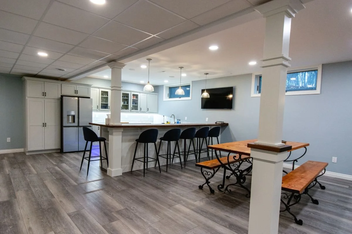 Modern basement with a kitchen, bar seating, and a dining area. The kitchen has white cabinets, a stainless steel refrigerator, and pendant lighting. The dining area has a wooden table with benches and decorative ironwork.