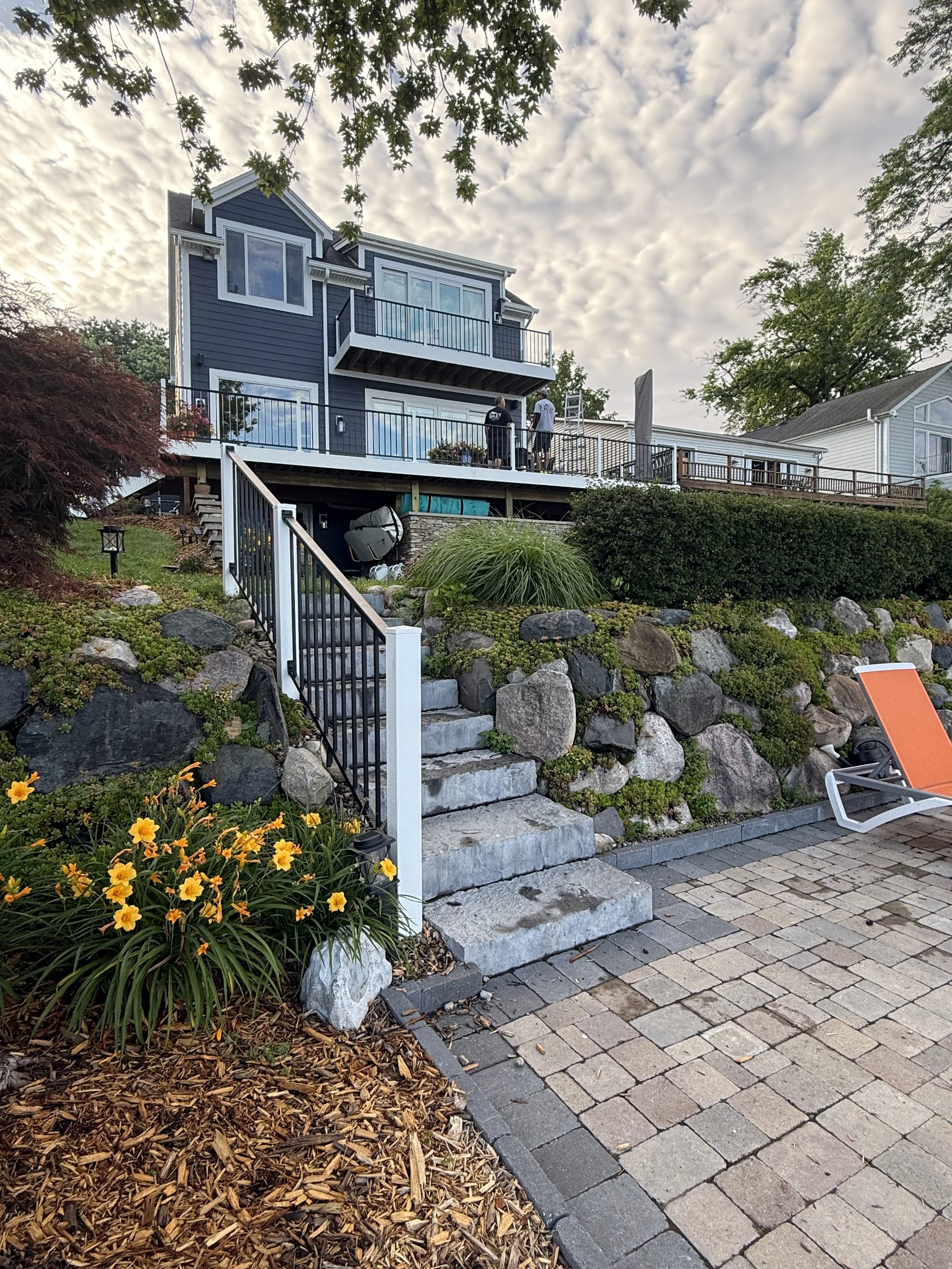 A backyard with a multi-level stone patio, a staircase, and a house with a deck. Two people are standing on the deck, and orange lounge chairs are on the patio. Flowers and greenery surround the area.