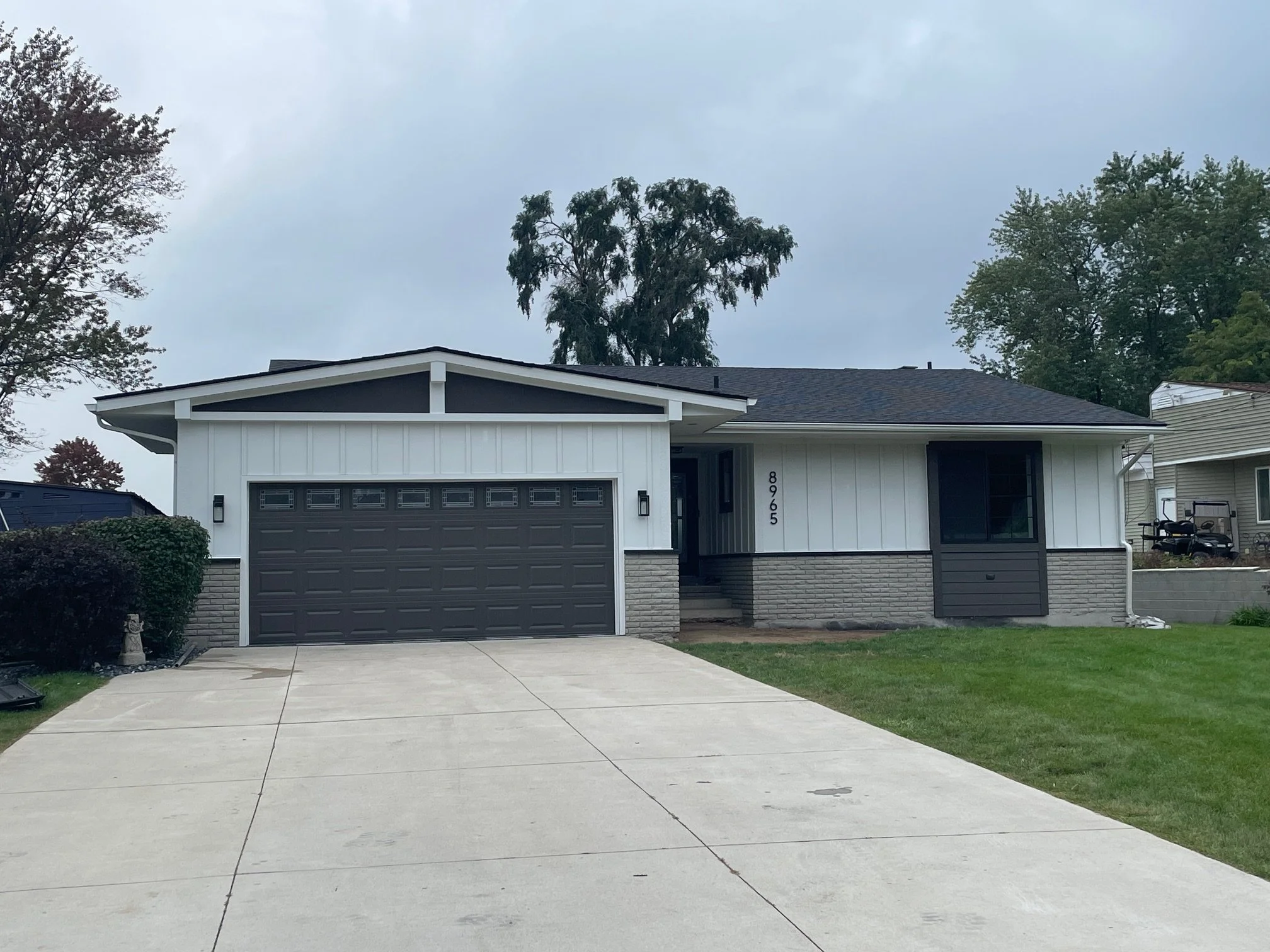A single-story house with white and dark gray exterior, a dark gray garage door, and a concrete driveway leading to the garage. The house has a small front porch and is surrounded by a lawn with trees in the background.