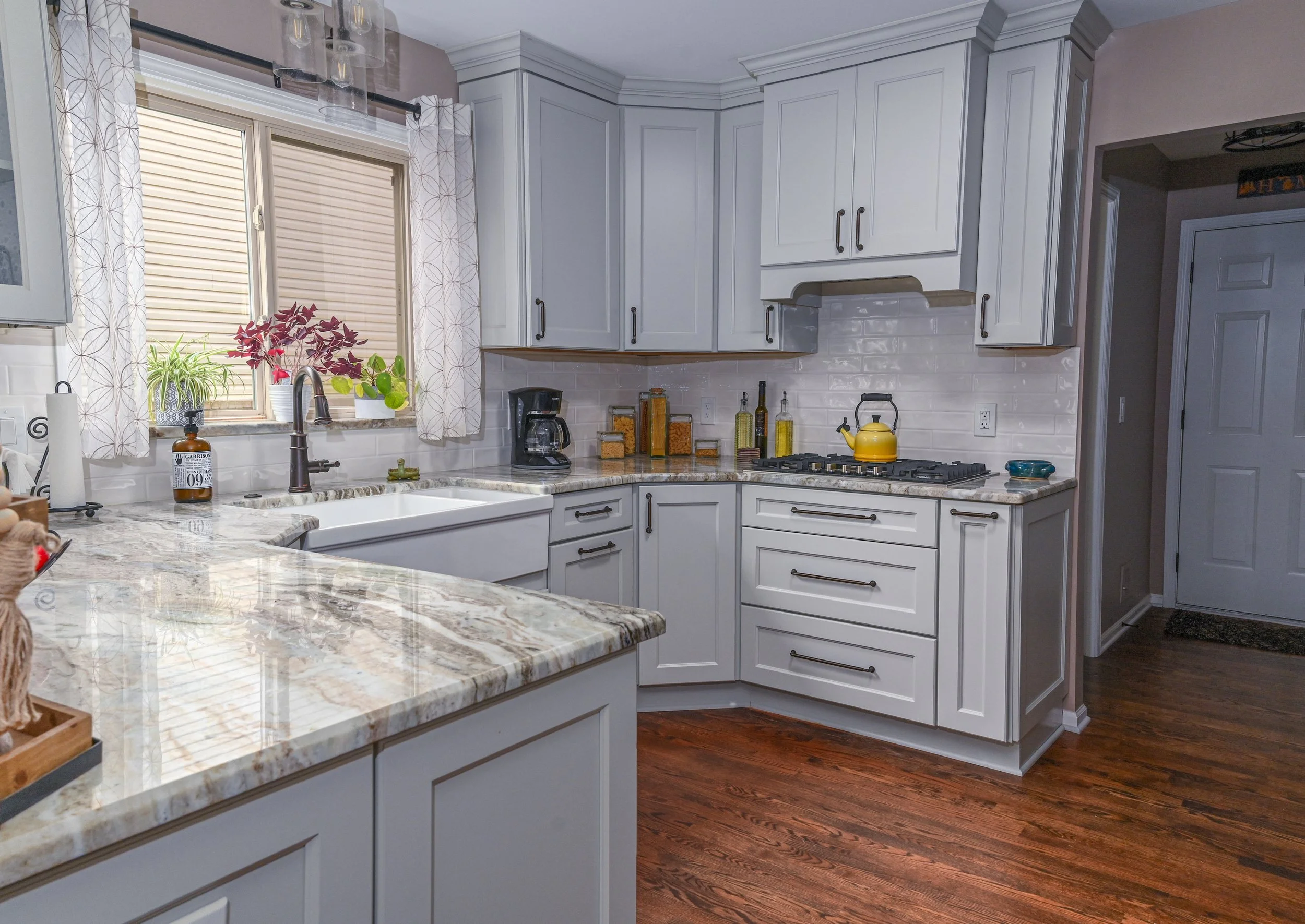 A kitchen with white cabinets, a granite countertop, a window with curtains, a coffee maker, a yellow teapot on a stovetop, and decorative jars on the counter. Dark hardwood floors are visible.