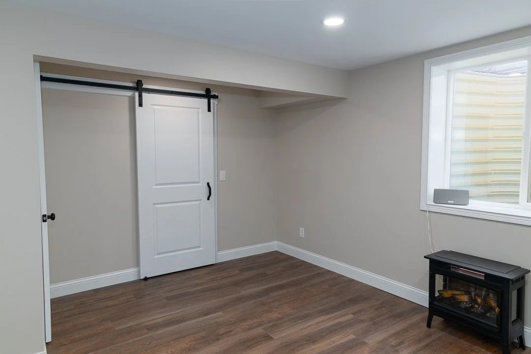 Empty room with beige walls, wooden floor, a sliding barn door, a window with blinds, a small electric heater, and a ceiling light.
