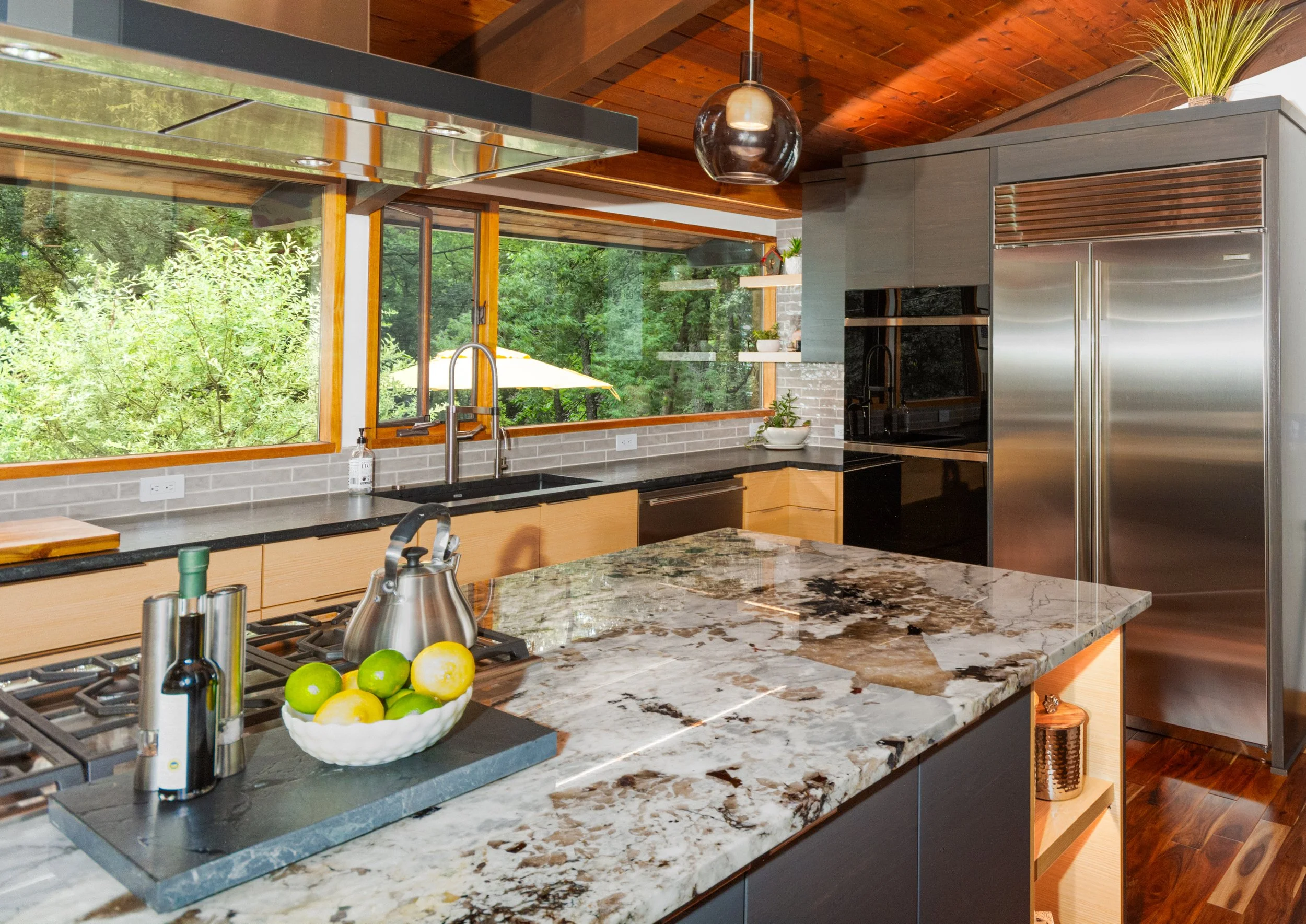 Modern kitchen with large windows overlooking greenery, marble countertop island, stainless steel appliances, and wooden ceiling.