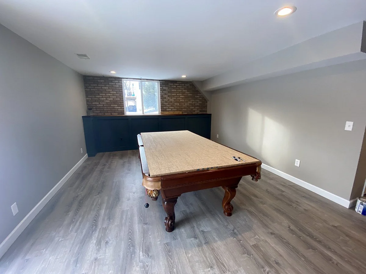 Empty basement room with a pool table, gray walls, a brick accent wall, a window, and ceiling lights.