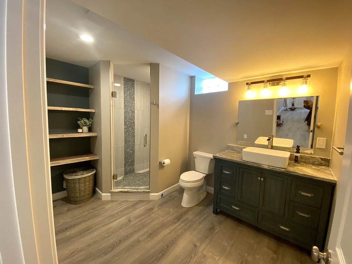 Bathroom with a gray vanity with a vessel sink, mirror, modern light fixtures, toilet, small glass-enclosed shower, open shelving with a plant and basket, and wood-like flooring.