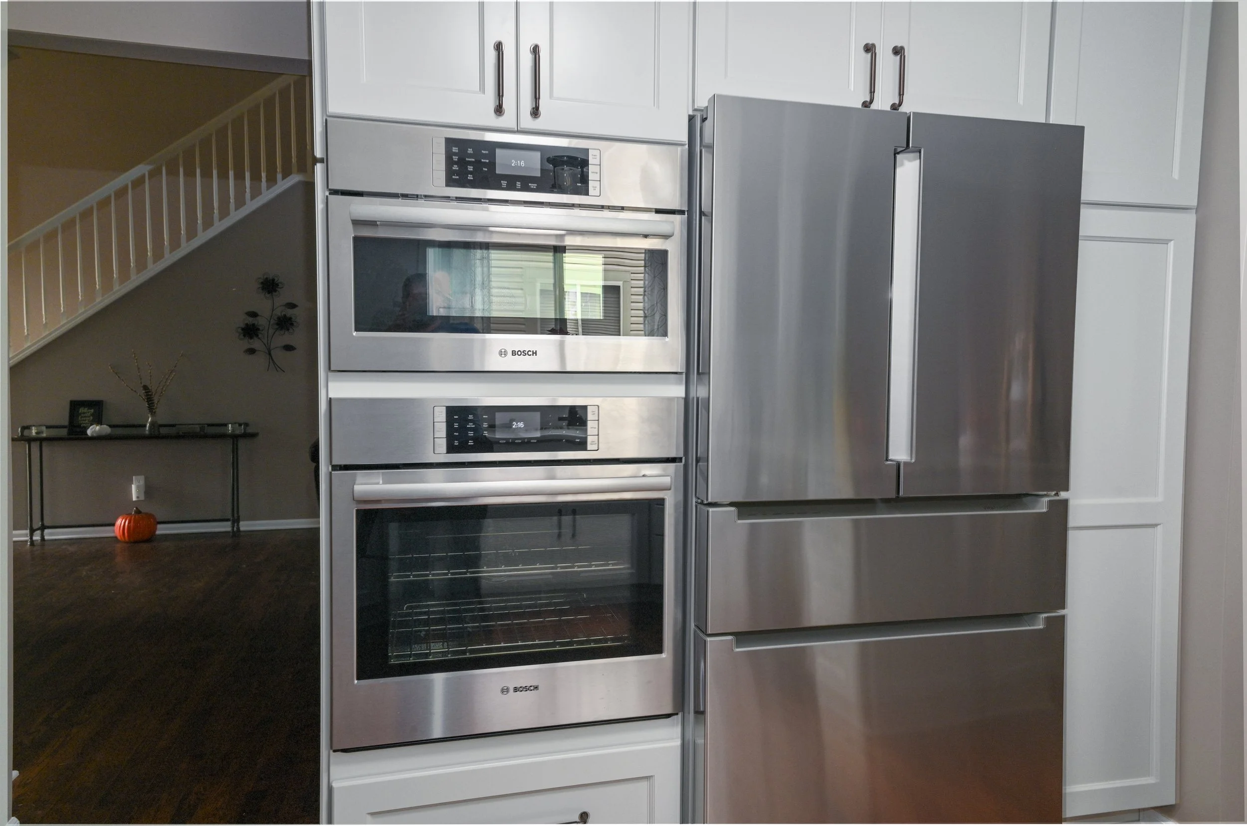 Stainless steel Bosch oven and microwave installed in a kitchen wall, with a side-by-side refrigerator to the right, white cabinetry above, and a view into a living area with dark wood floors and a stairway in the background.