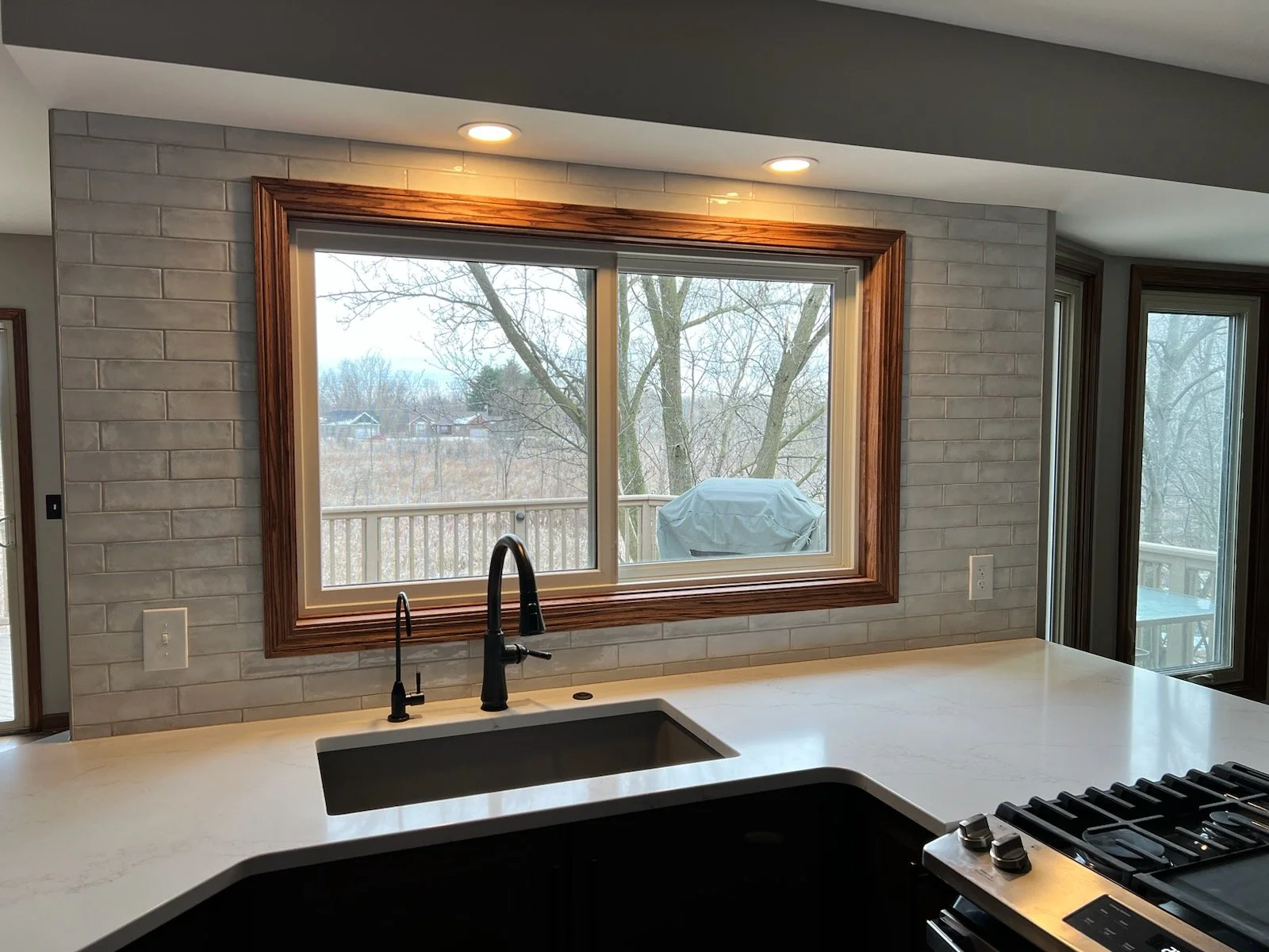 Modern kitchen with a large window overlooking a backyard with trees. Features include a white countertop, a black faucet, a sink, a gas stove, and recessed ceiling lights.