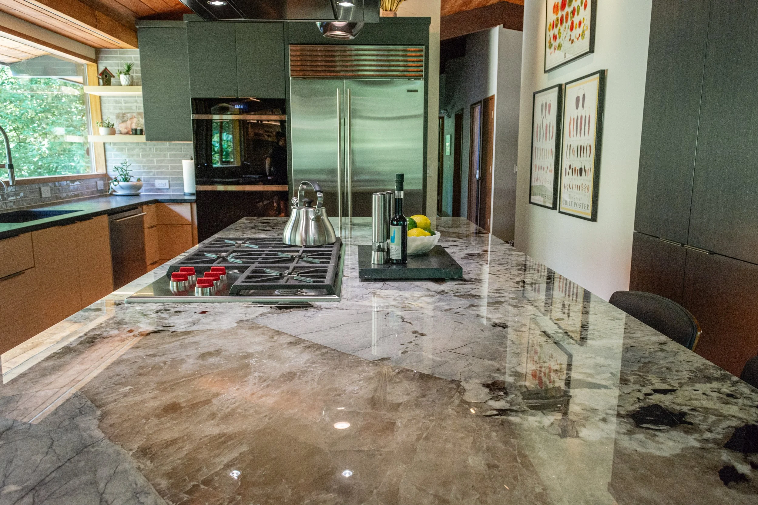 Modern kitchen with a large marble island containing a stovetop, tea kettle, and a bowl of lemons, with dark green cabinets, a stainless steel refrigerator, window, and framed artwork on the wall.