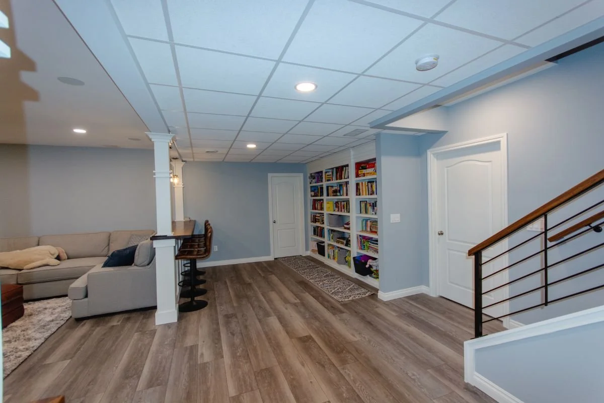 Living room with light blue walls, wooden floor, beige sectional sofa, built-in bookshelves filled with books, and staircase with wooden handrail and black metal spindles.