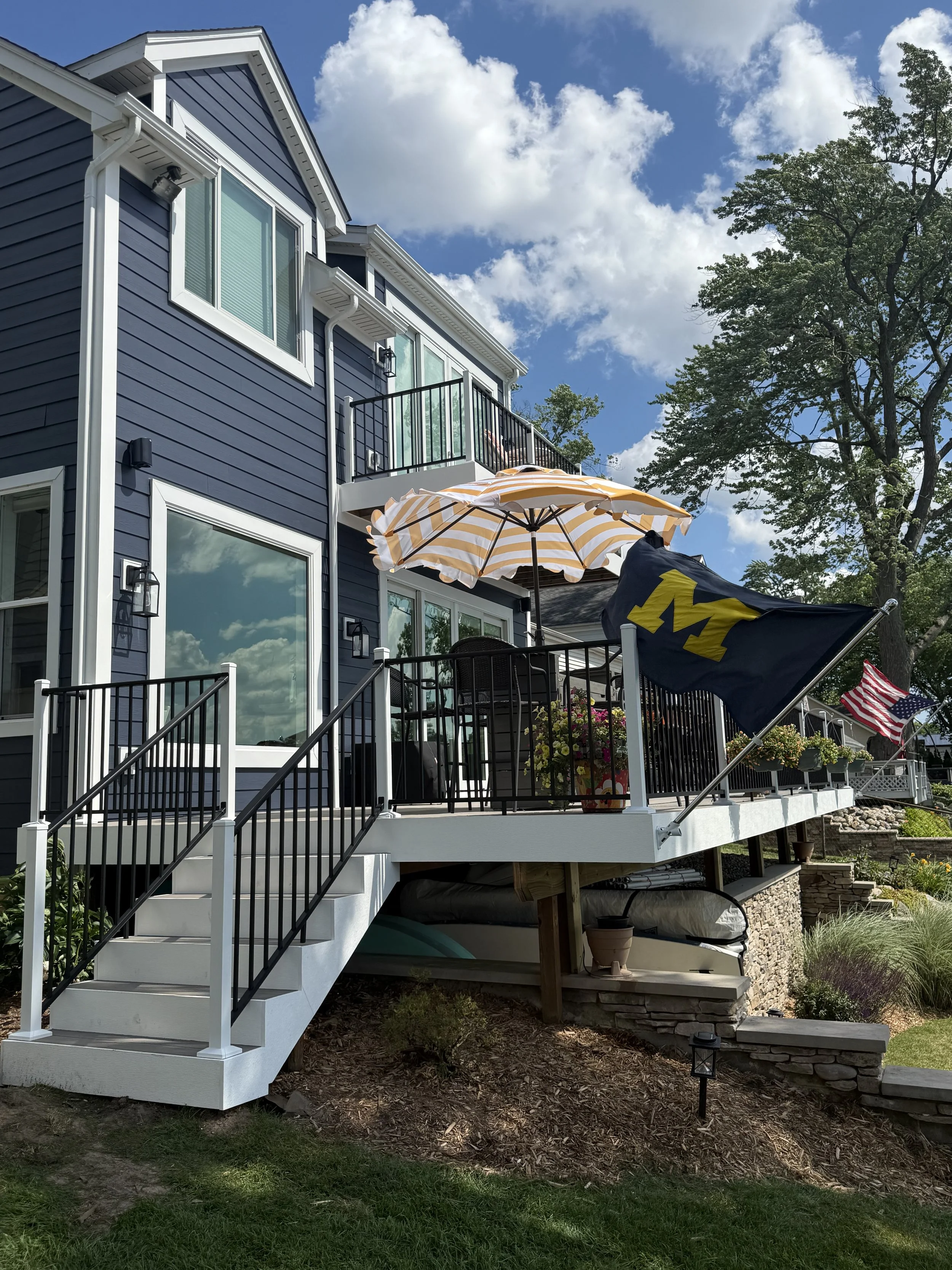 A two-story blue house with a white staircase leading to a backyard deck. The deck is decorated with umbrellas, potted plants, and flags, including a University of Michigan flag and an American flag. Tall trees and a partly cloudy sky are in the back