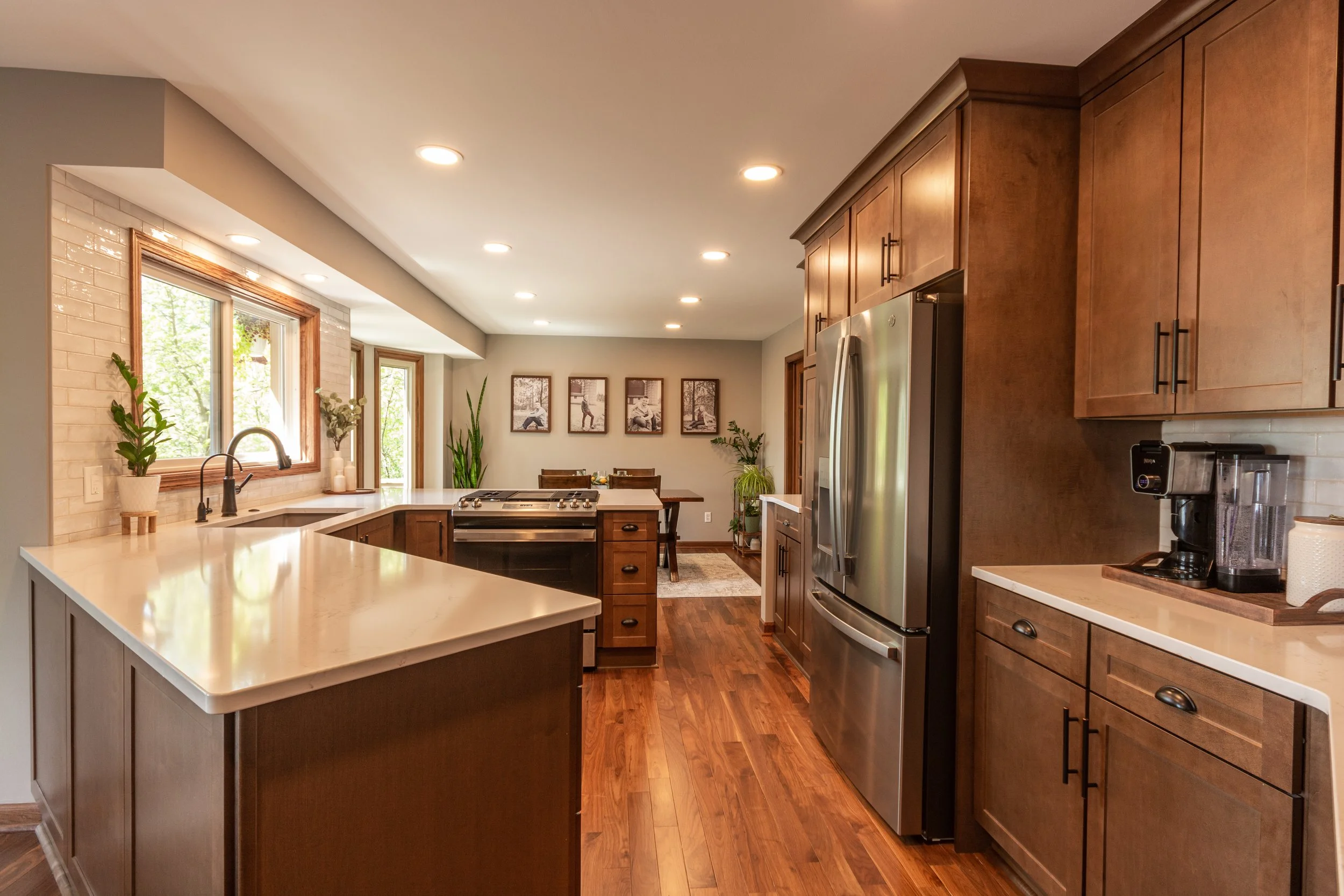 Kitchen with wood cabinets, stainless steel refrigerator, black stove, white countertops, and hardwood floor. There are three windows above the sink, plants on the counters, and a dining area in the background with framed pictures on the wall.