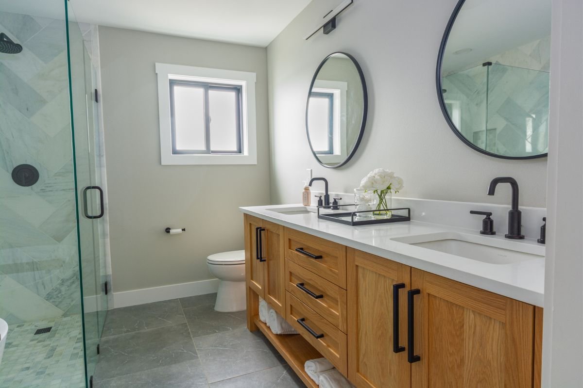 Modern bathroom with a wooden vanity, dual sinks, black faucet fixtures, two round mirrors, a glass shower enclosure, and a small window