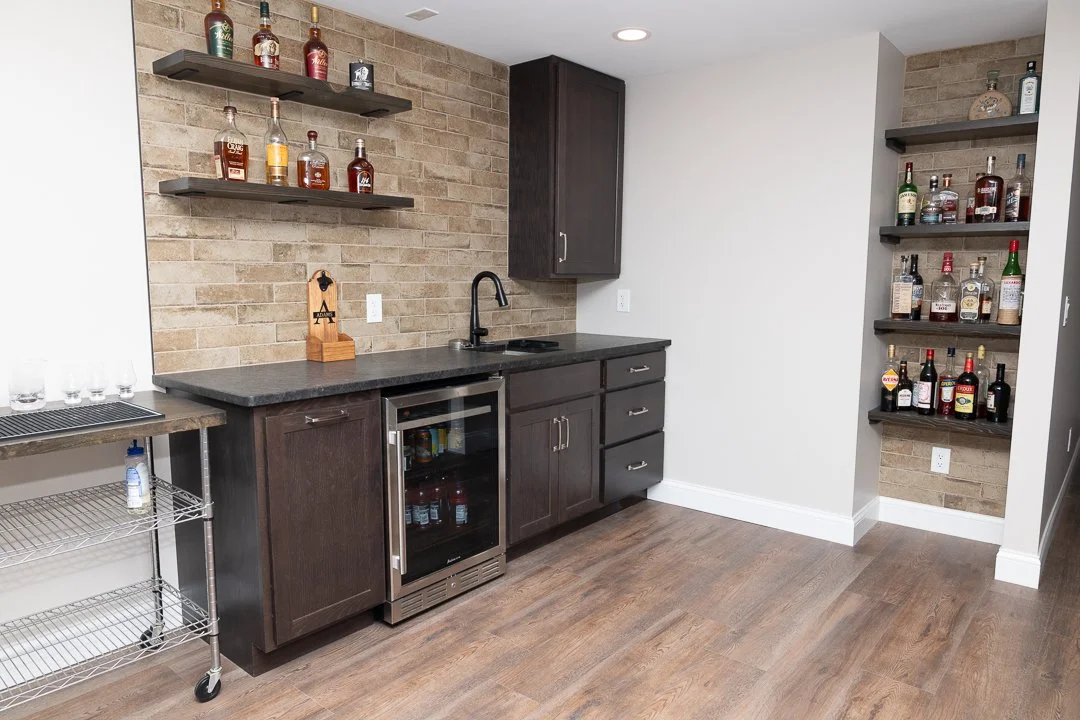 A home bar area with dark wood cabinets, a small wine cooler, open shelves with various bottles of liquor, a brick backsplash, and a countertop with a black faucet and sink, against a white wall with wood laminate flooring.