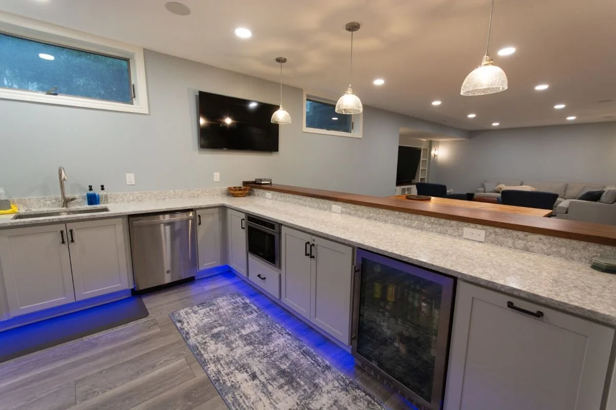 Modern basement kitchen with gray cabinets, granite countertops, stainless steel appliances, a mounted TV, pendant and recessed lighting, and a bar area with a wooden countertop.